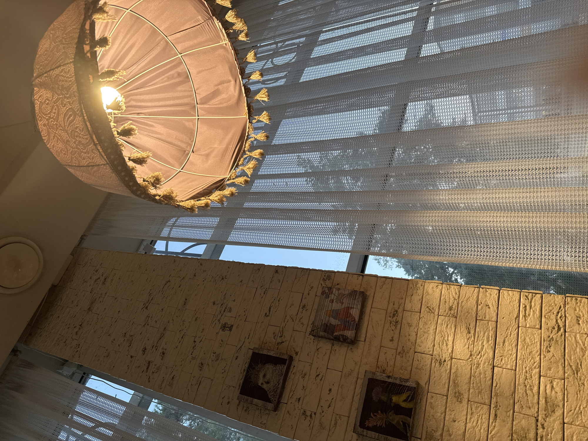 The image shows an interior view, likely a cafe or restaurant, with a focus on a decorative lampshade and a textured brick wall. The lampshade, made of a light-colored fabric with ornate detailing and fringed with yellow tassels, is prominently positioned in the upper right of the frame, illuminated by a warm light bulb. To the left, a light-colored brick wall is visible, adorned with three framed pictures. The bottom picture depicts two yellow birds with elaborate headpieces, the middle one shows three figures in colorful robes, and the top one features a bird-like creature with a feathered headdress. A small cross is mounted on the wall above the topmost picture. Sheer white curtains hang to the right, partially obscuring a large window that reveals a clear blue sky and the silhouette of trees. The overall lighting suggests it is daytime, with sunlight filtering through the curtains, casting patterned shadows. The location is identified as Abinsk, Russia.