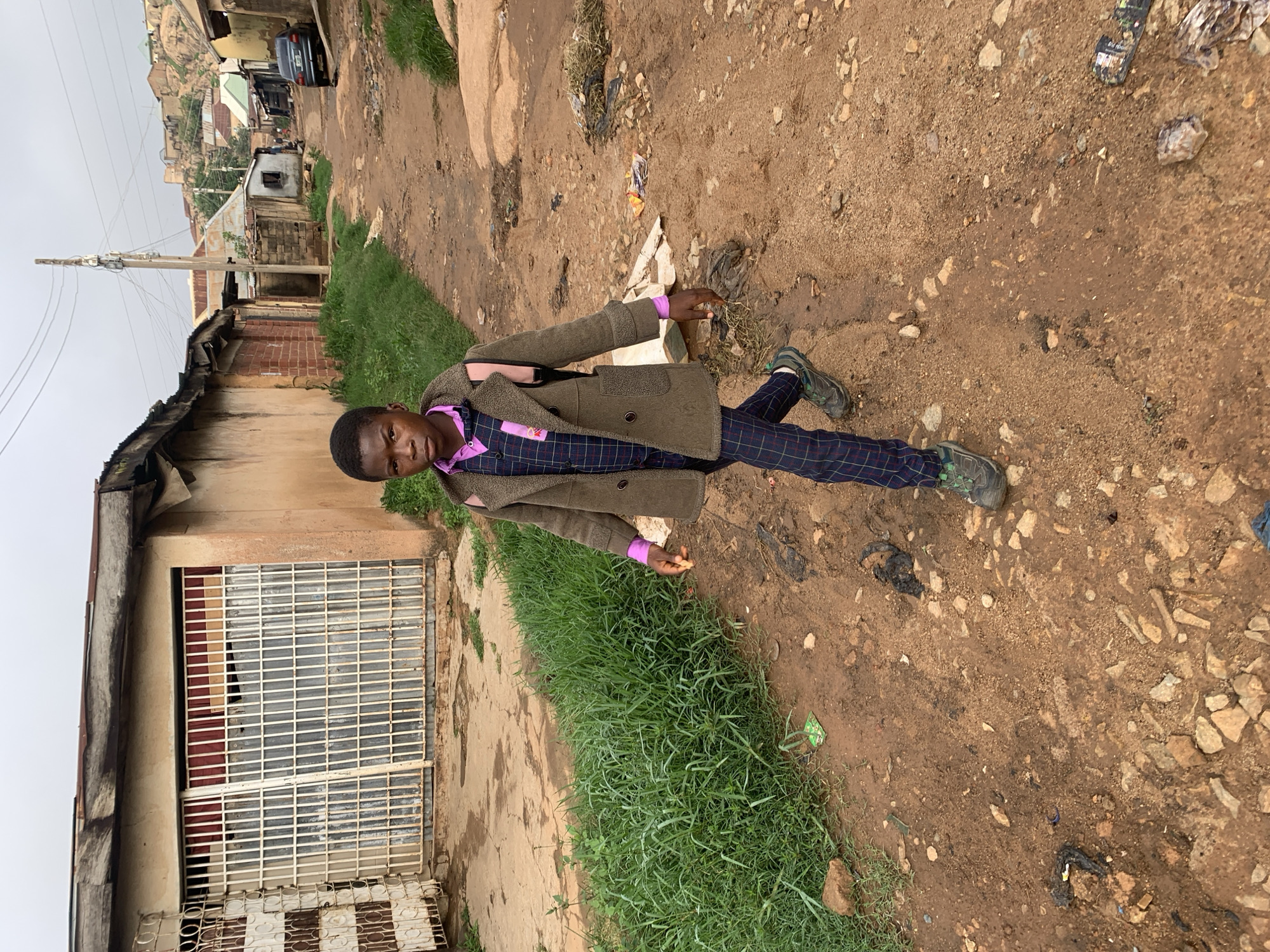 An outdoor, eye-level shot captures a young person standing on a rugged, unpaved path in what appears to be a semi-urban or rural area of Jos, Nigeria. The subject, a boy, is positioned slightly left of center, looking directly at the camera with a neutral expression. He wears a long-sleeved, olive-green coat over a vibrant purple collared shirt and dark plaid trousers. His shoes are green and grey, appearing practical for the terrain. His right hand is slightly extended forward, pointing towards some scattered debris on the ground.
The ground around him is a mix of reddish-brown dirt and numerous small to medium-sized rocks, with patches of green grass bordering the path on the left. Various pieces of litter, including plastic wrappers and dark, unidentifiable debris, are visible on the ground.
In the background, a row of modest, weathered buildings with corrugated iron roofs and grates over windows lines the left side of the path. A tall utility pole with numerous wires extends upwards towards a grey, overcast sky. Further back, a dirt road continues, with more buildings nestled against a backdrop of distant, rocky hills or mountains. A dark-colored car is partially visible on the road in the far distance. The scene is illuminated by soft, diffused daylight, suggesting an overcast day. No clearly legible text is visible on any objects or surfaces.