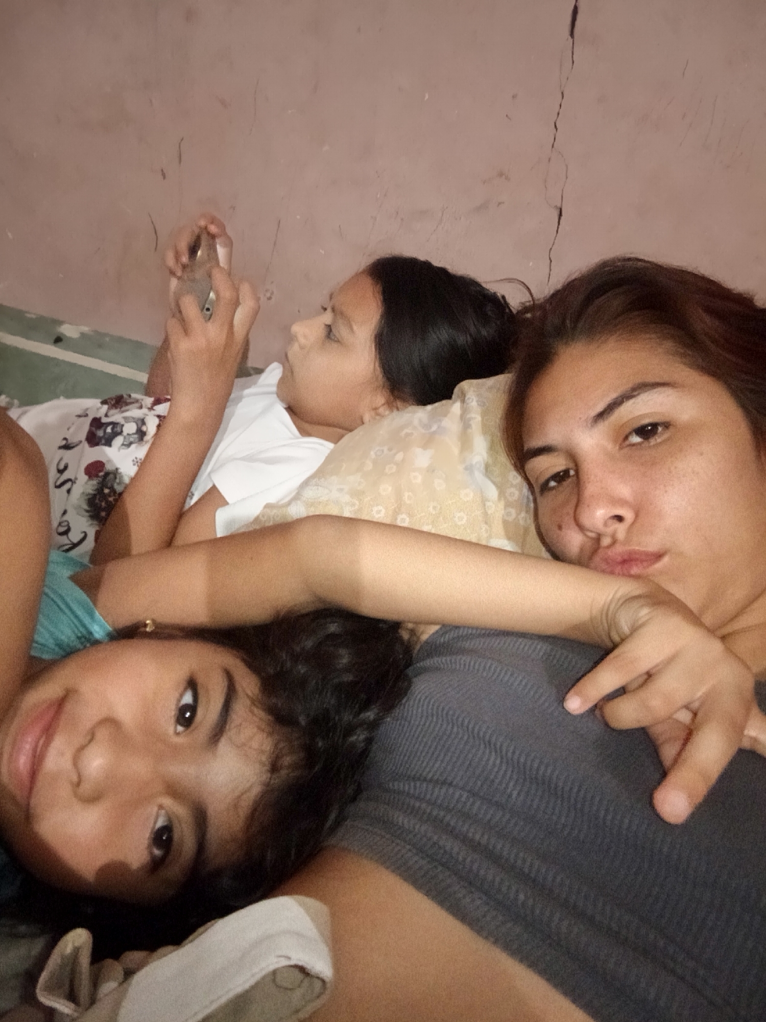 Three individuals are lying down indoors. A young girl is using a smartphone. Another young girl is smiling and looking at the camera. A woman is also looking at the camera with a slight pout. The background shows a plain wall with a crack. The location is Unknown, Unknown.