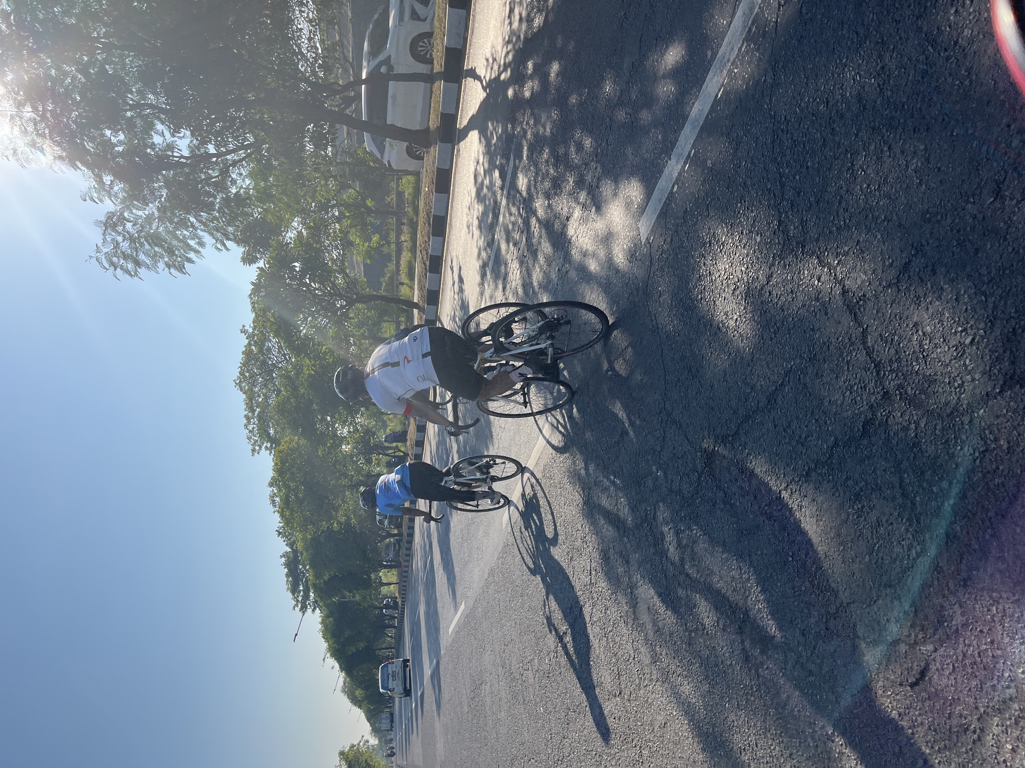 Two cyclists are riding on a paved road in Siring, Indonesia. The cyclists are positioned in the right lane of the road, and the sun is shining brightly, casting long shadows on the asphalt. The cyclist in front is wearing a blue shirt and black pants, while the cyclist behind is wearing a white shirt with red accents and black shorts. Both cyclists are wearing helmets. Trees line the side of the road, and there are cars parked on the shoulder. The sky is clear and blue. The image appears to be taken during the morning or late afternoon, given the angle of the sun and the length of the shadows.