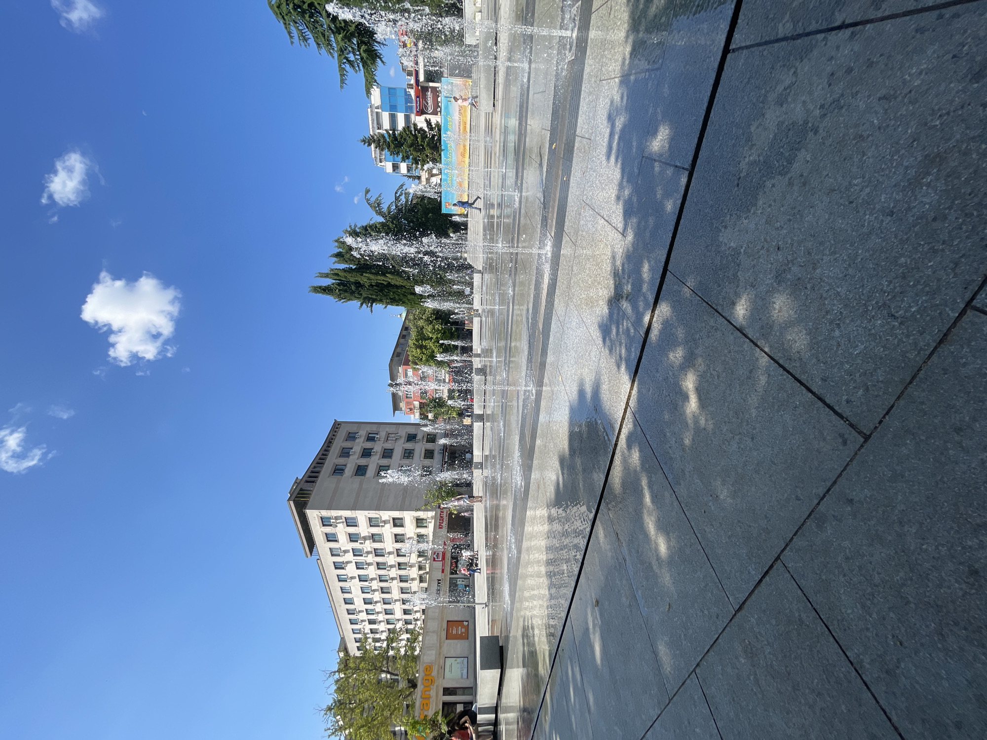 This is an eye-level, wide-angle, outdoor photograph of a public square in Stara Zagora, Bulgaria, featuring a modern water fountain. In the foreground, a large, paved area is wet from the water jets of a shallow fountain, with sunlight reflecting off the surface and casting dappled shadows from unseen trees. The fountain consists of numerous water spouts arranged in rows, creating a dynamic visual effect.
In the mid-ground, several people are present. On the left, three individuals are seated on a concrete bench. Further back, a few people walk through the fountain area. One person is walking to the right across the fountain, while another walks in the same direction in the background.
The background is dominated by urban architecture under a clear blue sky with a few scattered white clouds. To the left, a large, multi-story beige building with numerous windows and air conditioning units stands prominently. Adjacent to it is a smaller building with an orange facade featuring signage, possibly a shop or business. In the center and to the right, other buildings of varying heights and architectural styles are visible, including one with a blue glass facade. Tall, slender trees, likely cypress, are interspersed between the buildings. A large banner with text and imagery is displayed on one of the buildings in the background. The overall scene depicts a bright, sunny day in a lively urban public space.