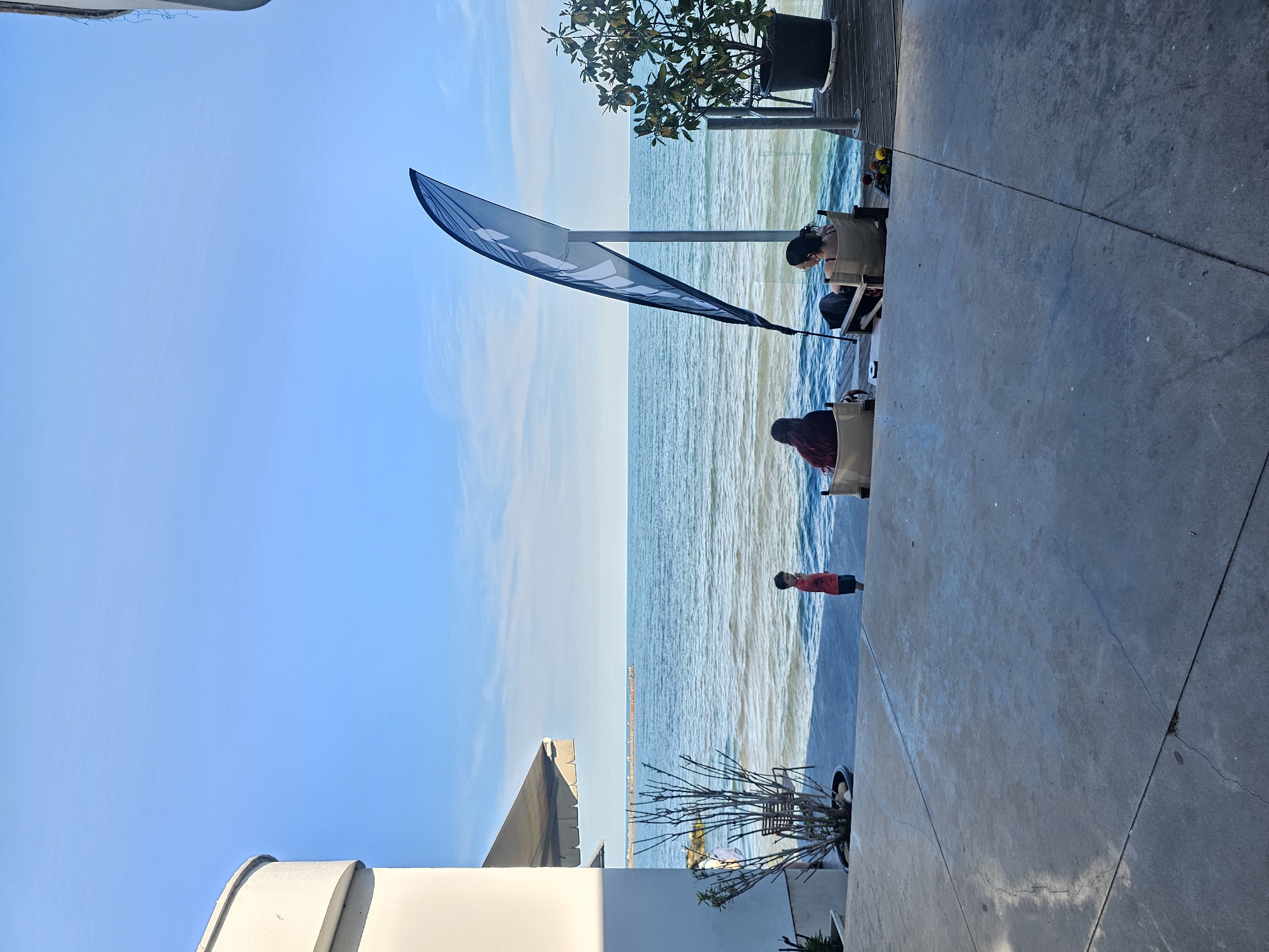 A wide shot captures an outdoor seating area by the sea under a clear blue sky. In the foreground, a concrete patio leads to a deck overlooking the water. Two individuals are seated in lounge chairs on the deck, facing the ocean. A tall, black and white flag with a curved design stands beside them. To the left, a small child in a red shirt stands on the patio, looking out at the sea. Further back, a pier stretches into the water, with a few people visible on it. The sea is calm with gentle waves, and a small boat is visible on the horizon. A potted plant sits on the deck to the right of the chairs. The scene suggests a relaxed atmosphere at a waterfront establishment.
