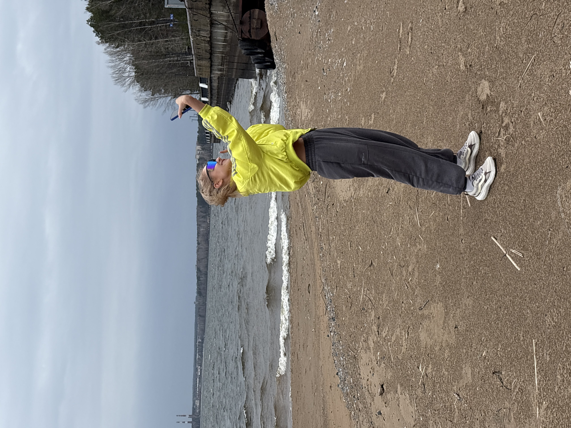 An image, rotated 90 degrees clockwise, captures a person standing on a sandy or dirt shoreline next to a body of water under an overcast sky. The person, with short, light-colored hair and wearing mirrored blue sunglasses, is dressed in a bright yellow track jacket featuring three white stripes along the outer sleeves, dark grey or black joggers with a small, illegible white text label on the left thigh, white socks, and grey and white athletic shoes.
The individual is actively engaged, holding a blue smartphone with their raised right hand, seemingly taking a photograph, video, or selfie of the scene. To their immediate left is the body of water, displaying small waves. The horizon reveals a distant landmass with bare trees and, further along, some industrial structures, possibly smokestacks. Behind the person, on the left side of the frame, there's a concrete or stone wall, possibly part of a pier or embankment, with darker, industrial-looking fittings. The foreground consists of a coarse, sandy-brown path or beach area with sparse dry vegetation and small pebbles. The weather appears cool and cloudy, indicating daytime without direct sunlight. The scene suggests a lakeside or riverine environment, potentially near an industrial area, consistent with the context of Chaikovsky, Russia.