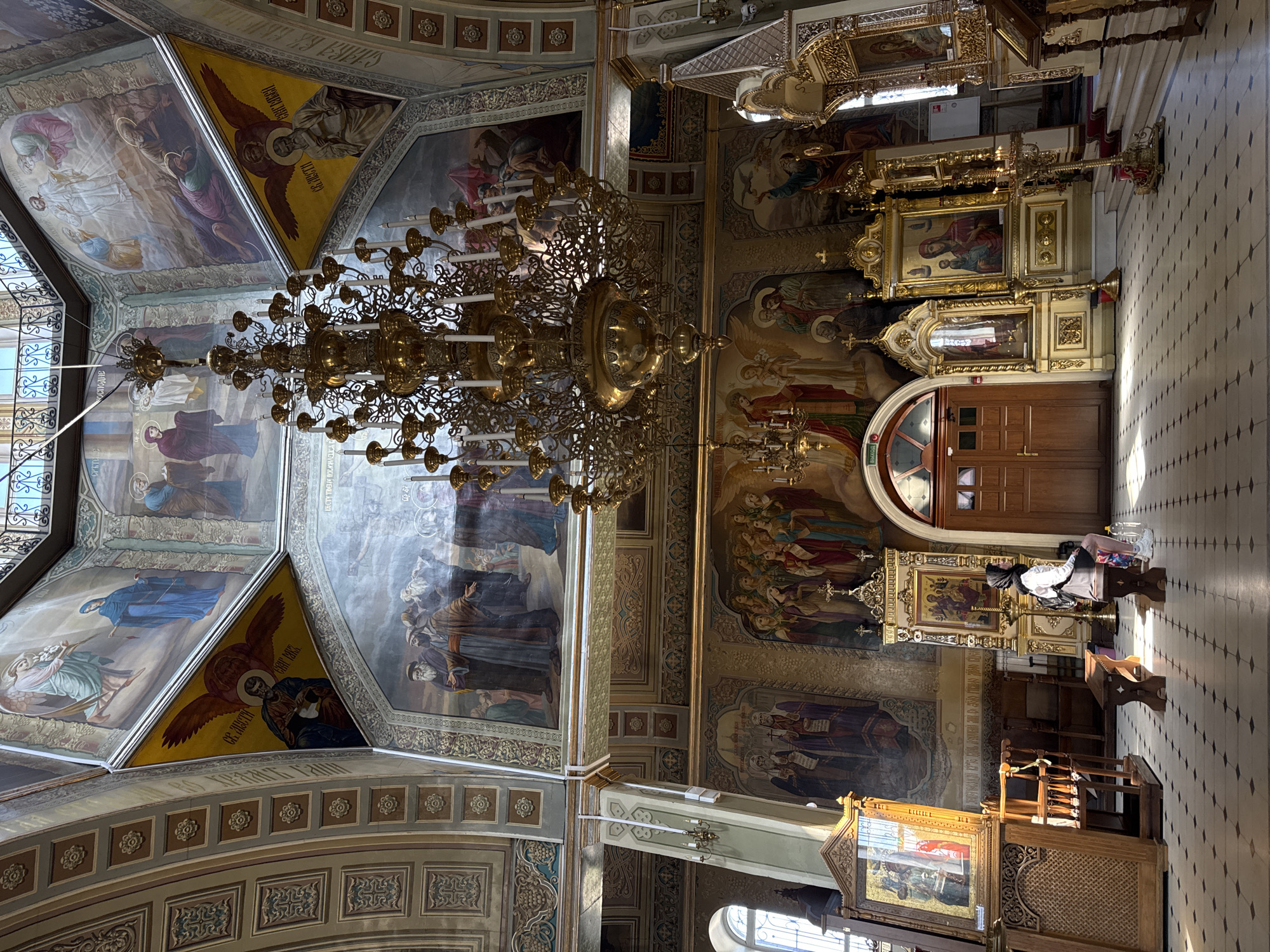 An interior view of an ornately decorated church features a grand chandelier hanging from the domed ceiling. The walls and ceiling are adorned with religious murals depicting biblical scenes and figures. Sunlight streams through windows, casting shadows on the tiled floor. A woman wearing a hijab and white jacket is seated on a bench, facing a golden iconostasis. To her left, a large icon is displayed on a wooden stand. The church is empty except for the seated woman.