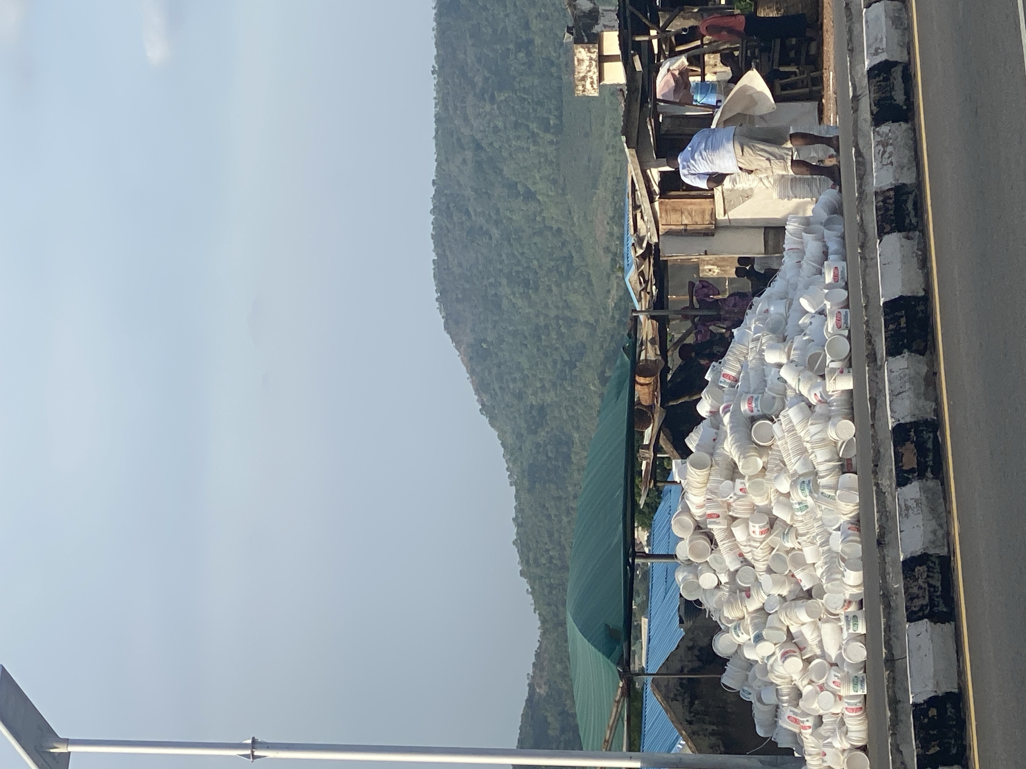 This image depicts an outdoor scene, likely a commercial or storage area, set against a backdrop of a large, verdant mountain under a clear, bright sky. The foreground is dominated by a substantial mound of white plastic cups or containers, stacked loosely. Many of these containers have faint colored labels or markings, although specific text is not legible.
Several individuals are present. One person, dressed in white attire resembling a kurta and light trousers, stands near the top of the pile of containers, appearing to be engaged in activity or observation. Another person, wearing a darker, possibly red, top, is visible further back within a shaded structure. The area includes simple structures or stalls, some partially covered by a green tarp and others by a blue corrugated roof.
To the right of the piled containers, a vertical barrier with alternating black and white stripes, possibly a roadside marker or boundary, runs along a paved surface that suggests a road or pathway. The clear sky and strong illumination indicate it is daytime. The scene conveys a sense of commerce or material handling, possibly related to a market stall or recycling operation in a semi-urban or rural setting like Zango-ndaji, Nigeria.