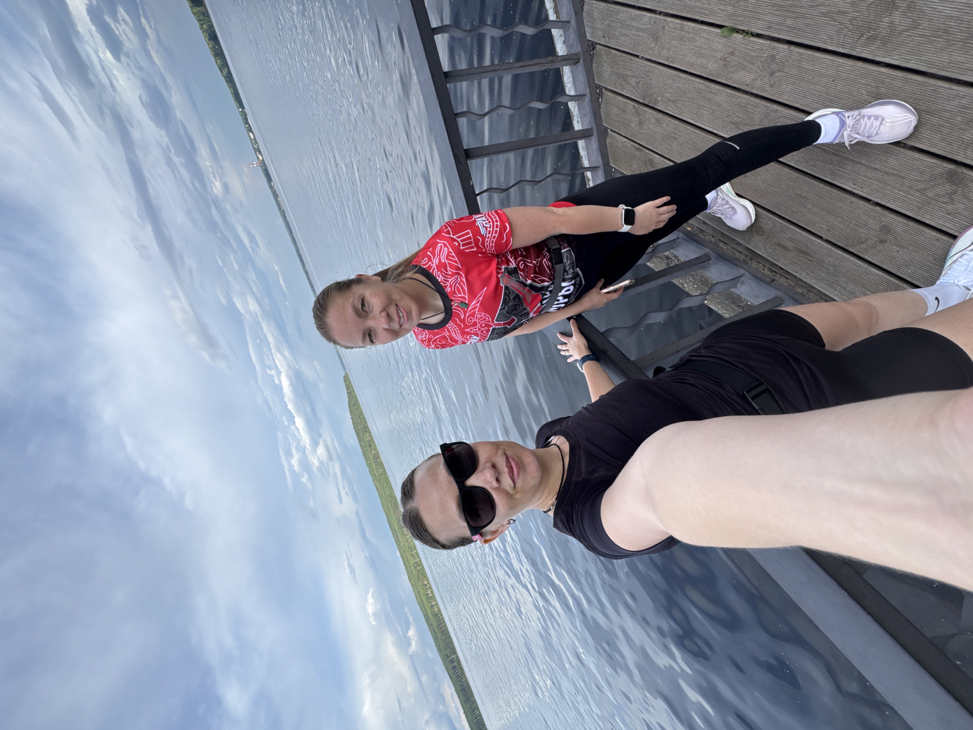 The media file depicts two women on a wooden pier overlooking a body of water. The woman in the foreground is wearing sunglasses, a black t-shirt, and black shorts. She is holding a phone to take a selfie. The woman in the background is wearing a red t-shirt with a graphic design, black leggings, and white sneakers. She is also looking towards the camera and smiling.
The scene appears to be outdoors, with a cloudy sky overhead and a calm body of water in the background. The pier is made of wooden planks and has a metal railing. The overall mood is casual and relaxed, suggesting a leisure activity or a break during an outdoor excursion. The location cues, such as the specific type of pier and the surrounding landscape which includes a line of trees on the distant shore, are consistent with a location like Chaikovsky, Russia. The time of day appears to be daytime, given the lighting conditions.
There is visible text on the red t-shirt of the woman in the background. The text is white and appears to be in Cyrillic script. It is difficult to read clearly from the provided image, but it seems to be part of a graphic design or slogan. No other significant text is visible in the image.