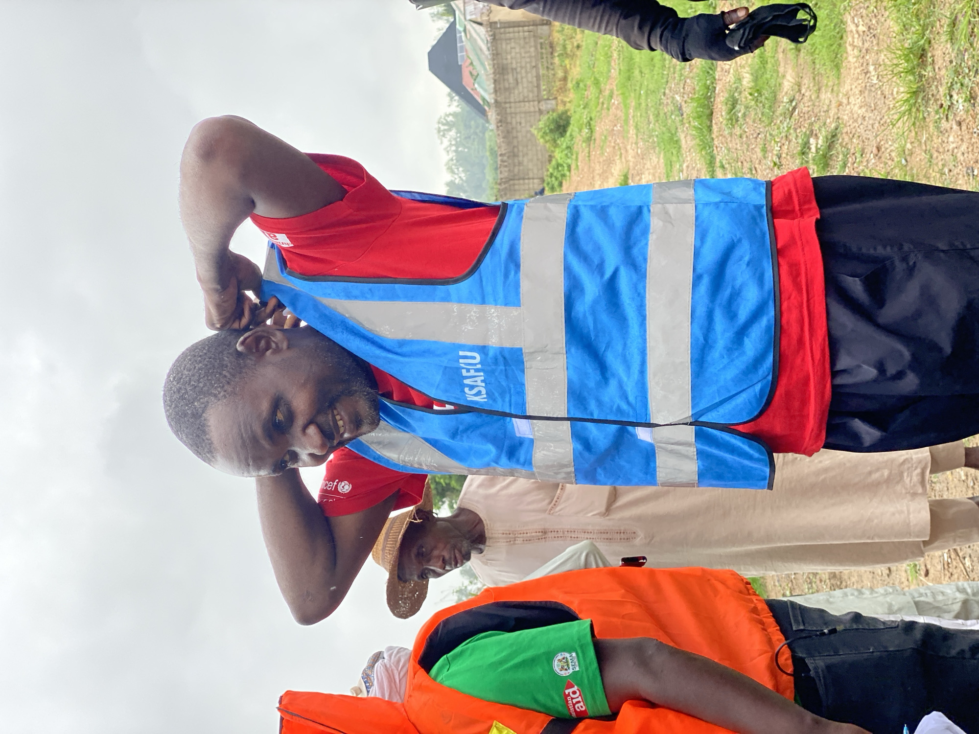 The image shows a medium shot of a Black man in a red t-shirt and a blue high-visibility vest with reflective silver stripes, adjusting the vest over his head. He has short, dark hair and a beard and is smiling. Behind him, slightly out of focus, are at least two other people. To the left, a person in an orange high-visibility vest with a green shirt underneath is visible from the shoulders down. To the center, a man wearing a straw hat and a light-colored traditional garment is visible. The background shows a grassy area with a fence and some buildings with dark roofs. The sky is overcast. The image appears to be taken outdoors in an urban or semi-urban environment, possibly in Kawo, Nigeria, given the context.