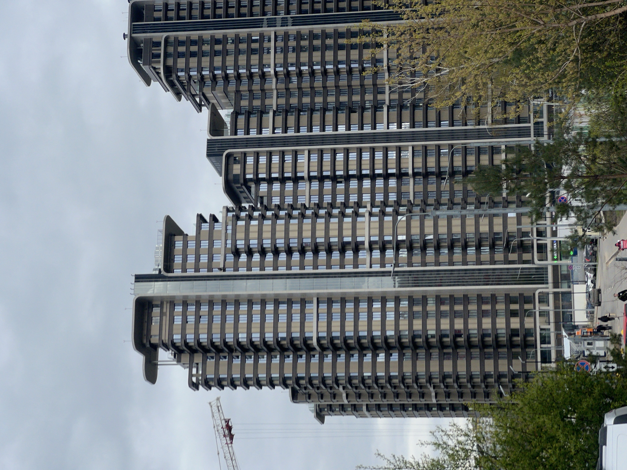 The image shows a view of the residential complex "Gorki Gorod" in the city of Khimki, Russia. The complex is made up of several high-rise buildings with a unique architectural style, featuring projecting ledges and dark vertical elements on the facade. The buildings are tall and imposing, taking up a large part of the frame.
In the foreground, there are trees, indicating that the complex is situated in an urban area with some greenery. There is also a crane visible, suggesting ongoing construction or maintenance. In the background, there is a street with cars and some pedestrians, indicating the location of the complex within the city. The sky is overcast, suggesting a cloudy day.