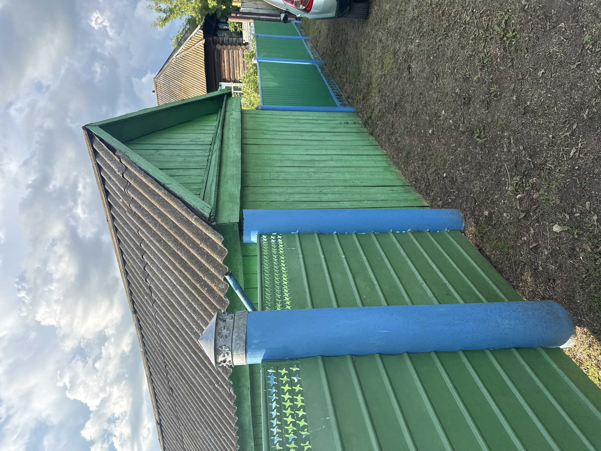 In the village of Chuy-Ataevo, Russia, a weathered, grey, corrugated roof caps a rustic green wooden structure. A blue cylindrical post, painted in a bright, matte finish, supports the structure and a section of green wooden fence. This fence, constructed from horizontal planks, is adorned with a decorative border of light green and blue star-like patterns along its top edge. Beyond this fence, a secondary green corrugated metal fence with blue posts stretches into the distance, bordering a grassy area. In the background, another wooden building with a similar grey roof is visible, along with lush green foliage. The sky above is a dramatic mix of light and dark grey clouds. The ground in the foreground is a mixture of sparse grass and dark soil. A portion of a silver car is visible on the far right of the image.
