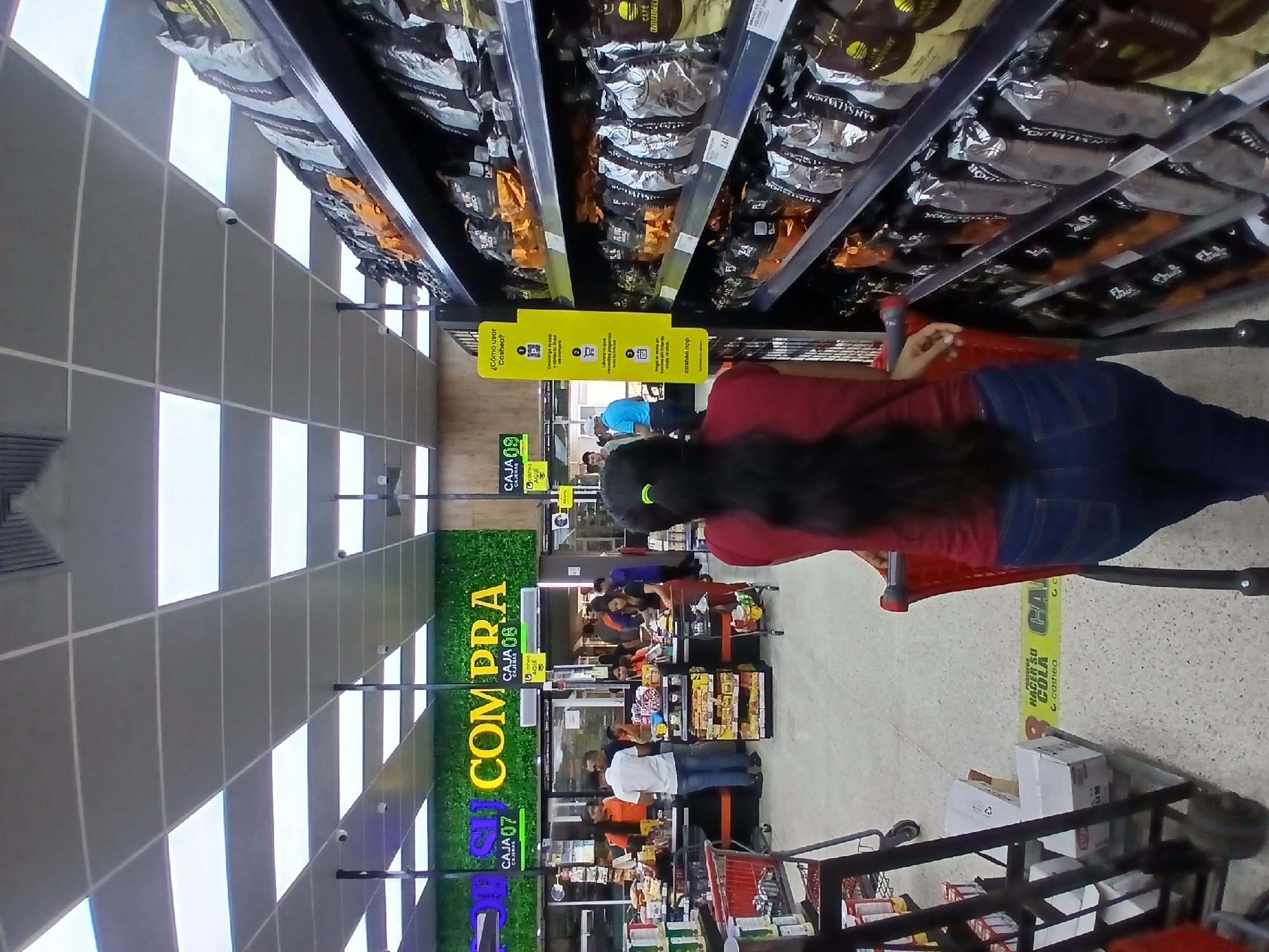 The image depicts the interior of a supermarket in Ciudad Guayana, Venezuela. A woman wearing a red shirt and blue jeans is standing in front of a shopping cart, positioned near the checkout lanes. The checkout lanes are numbered "Caja 07," "Caja 08," and "Caja 09." Signage indicates that the store accepts payments via "Cashea," an app which allows customers to pay in installments. Shelves stocked with packaged goods, including "San Salvador" and "Cafe Amanecer," are visible above the woman and along the edges of the image. Other people are present in the checkout area. The environment includes tiled floors and a drop ceiling.