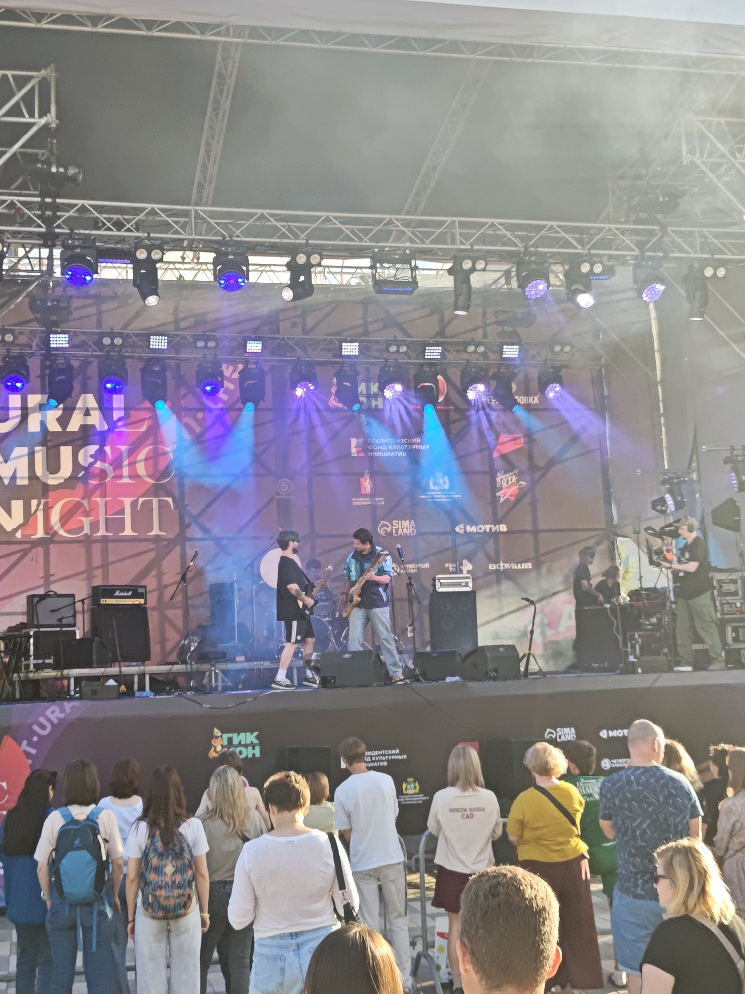 The image depicts an outdoor music festival in Yekaterinburg, Russia, likely during the daytime given the bright sunlight casting shadows and the visibility of the sky. A band is performing on a stage equipped with professional lighting and sound equipment. Two musicians are visible on stage: one playing a guitar and singing, wearing a black t-shirt and shorts, and another playing a guitar, wearing a blue and white checkered shirt. Several other band members and technical crew are positioned behind them, engaged in their roles.
In front of the stage, a crowd of spectators is gathered, facing the performers. People are standing and watching the concert, some with their backs to the camera. The audience appears to be a mix of ages and genders, with various clothing styles suggesting a casual event. Some individuals are holding backpacks or bags.
The stage backdrop features a large banner with text that appears to be in Russian, including "URAL MUSIC NIGHT" in English. Various sponsor logos and names are also displayed on the backdrop. The overall environment suggests a lively and public event, with a focus on live musical entertainment. The presence of professional camera equipment on the right side of the image indicates that the event is being filmed or broadcast. The festival seems to be taking place in an urban setting, likely a park or public square, as suggested by the presence of a fence separating the audience from the stage area.