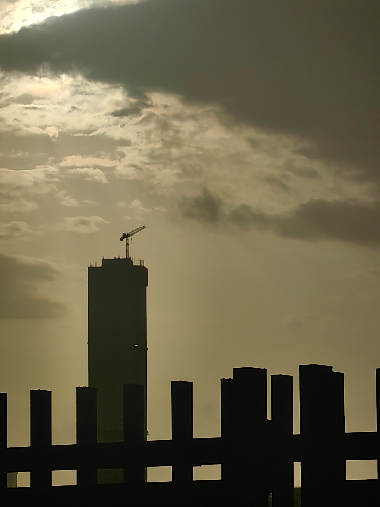 A silhouetted view of a cityscape under a cloudy sky. The primary subject is a tall, dark, rectangular building under construction, topped with a crane. In the foreground, a dark, serrated silhouette of a railing or fence runs horizontally across the lower portion of the image. Behind this fence, various other silhouetted structures of varying heights are visible, suggesting a dense urban environment. The sky is overcast with a mix of bright, diffused light and darker, dramatic clouds, creating a subdued and atmospheric lighting condition. The overall impression is that of a city at dawn or dusk, with a focus on the contrast between the man-made structures and the natural light. The image is consistent with a view of the Mumbai skyline.