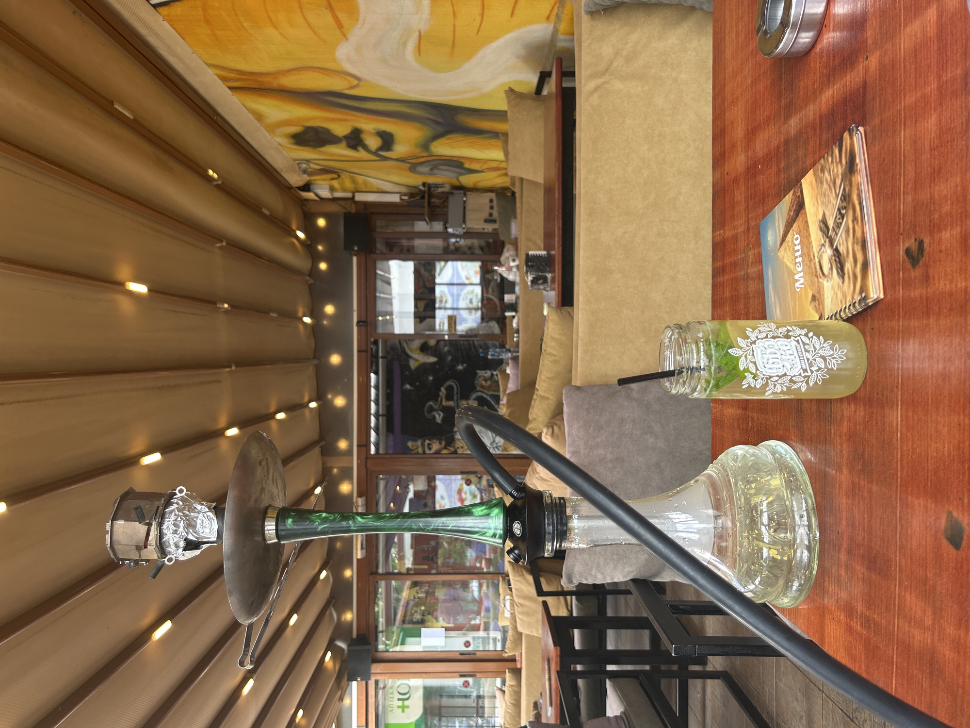 A hookah sits on a wooden table in a cafe. Next to it is a glass mason jar with a drink and a menu. The cafe has a mural on the ceiling and is located in Unknown, Unknown.