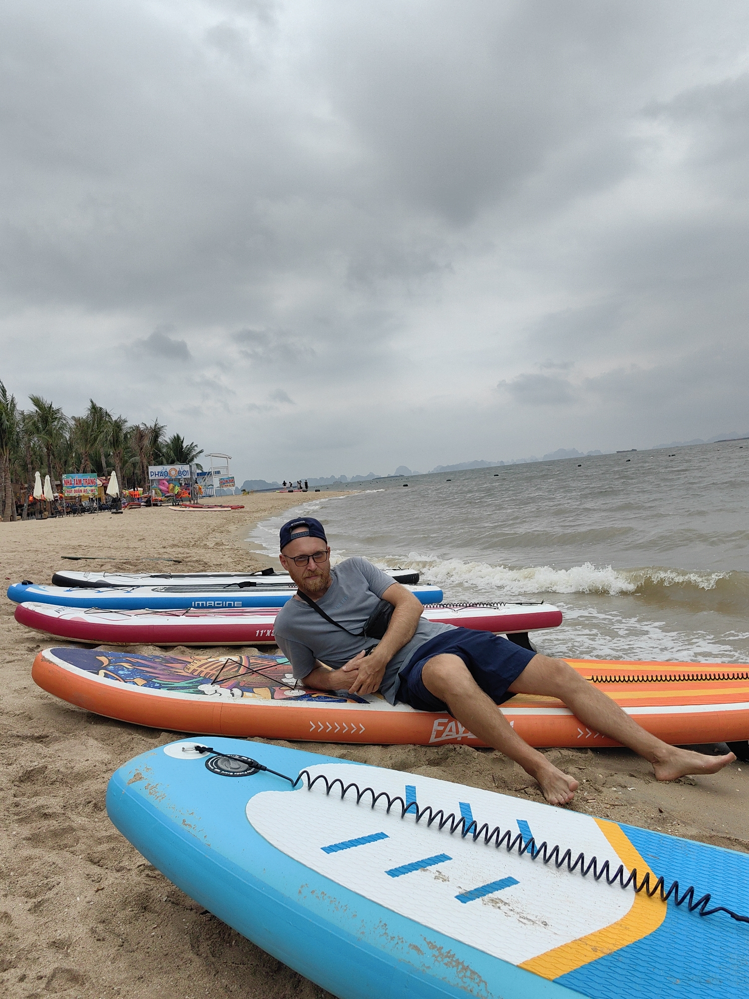 This beach scene in Bai Chay Ward, Vietnam, captures a relaxed atmosphere under an overcast sky. In the foreground, a man with a beard and glasses, wearing a grey t-shirt, dark blue shorts, and a dark baseball cap worn backward, is lying on his stomach, propped up on his elbows. He is resting on an orange inflatable stand-up paddleboard (SUP) marked "FALCO". Several other colorful paddleboards, including blue, red, and light blue ones, are arranged on the sandy beach around him. Brands like "IMAGINE" and dimensions "11'X32"X6"" are visible on some of the boards, which also feature coiled leashes.
The sandy beach extends into the background, lined with numerous palm trees. Beach umbrellas, chairs, and several signs with Vietnamese text are visible among the trees, suggesting a resort or public beach area. Text on signs includes "NHA TAM-TRANG" and "PHAO BƠI". A lifeguard stand can also be seen in the distance. The ocean, with small waves breaking on the shore, occupies the right side of the frame. Buoys are scattered across the water, and distant land masses or mountains are visible on the horizon beneath the grey, cloudy sky. The overall impression is one of leisure, with the man appearing calm and relaxed, possibly resting before or after a paddleboarding session.