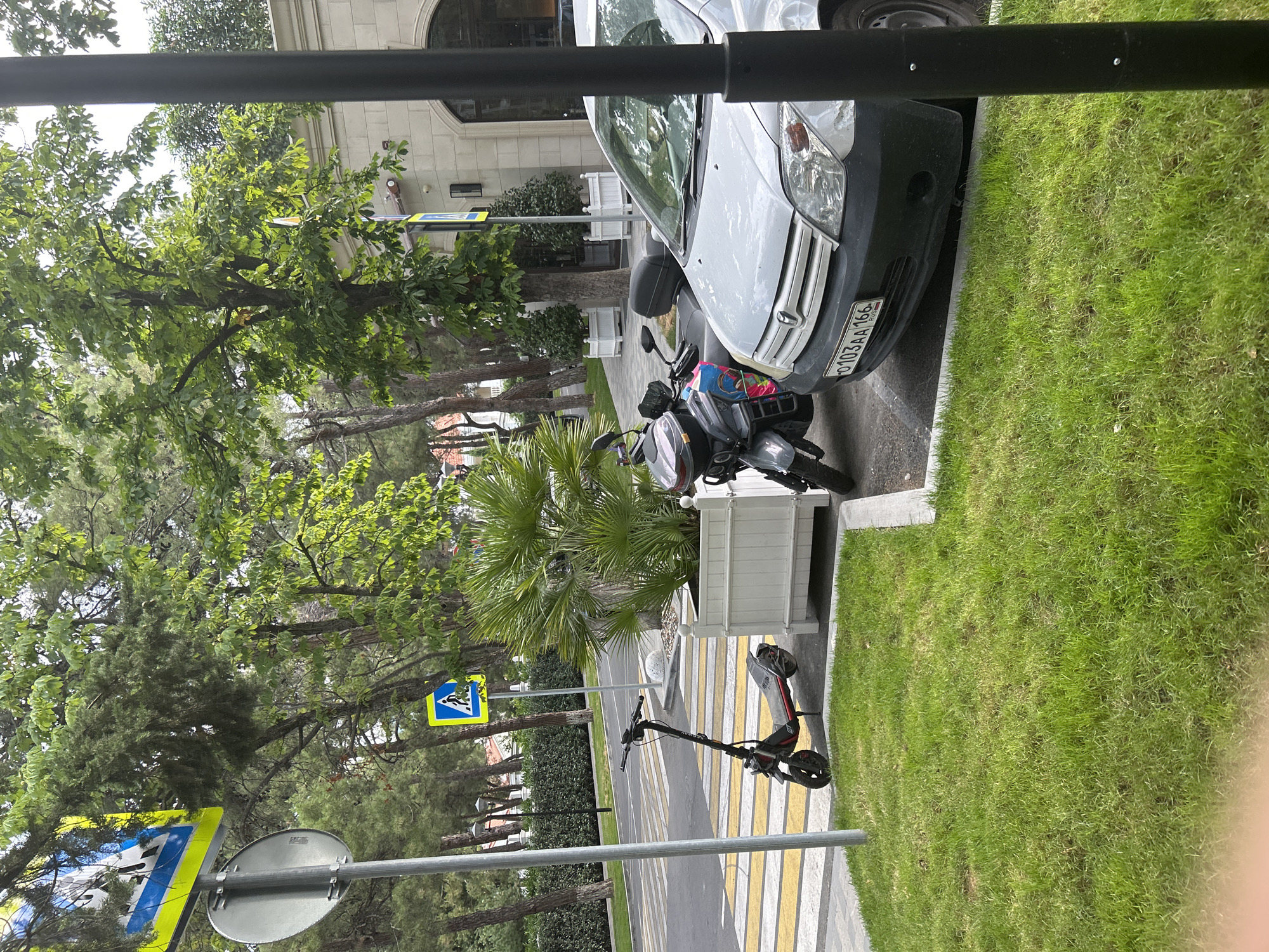 A street scene in Gelendzhik, Russia, features a pedestrian crossing with yellow and white stripes on asphalt. A silver car is parked to the right, its license plate reading "0103AA 166 RUS." Next to the car, a grey scooter with a colorful graphic on its side is parked. In the background, there are trees and a building with a light-colored facade. A pedestrian crossing sign is visible on a pole. A black metal pole is in the foreground on the right.