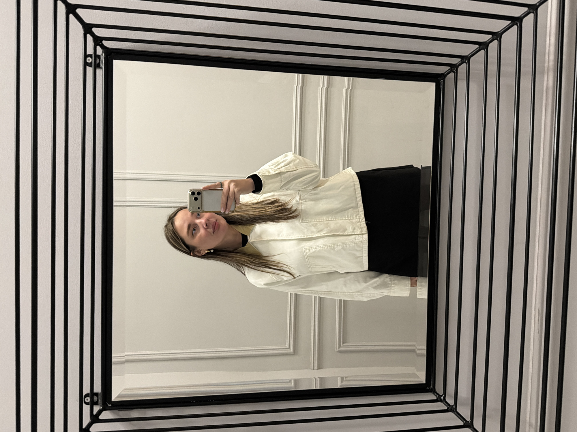 A person is reflected in a mirror. The mirror has a black frame with a geometric, cage-like structure around the edge. The person is holding a smartphone to take a picture. The person appears to be wearing a black top and a white jacket. The mirror reflects a white wall with decorative molding. The scene does not explicitly contain details allowing identification of Moscow, Russia.