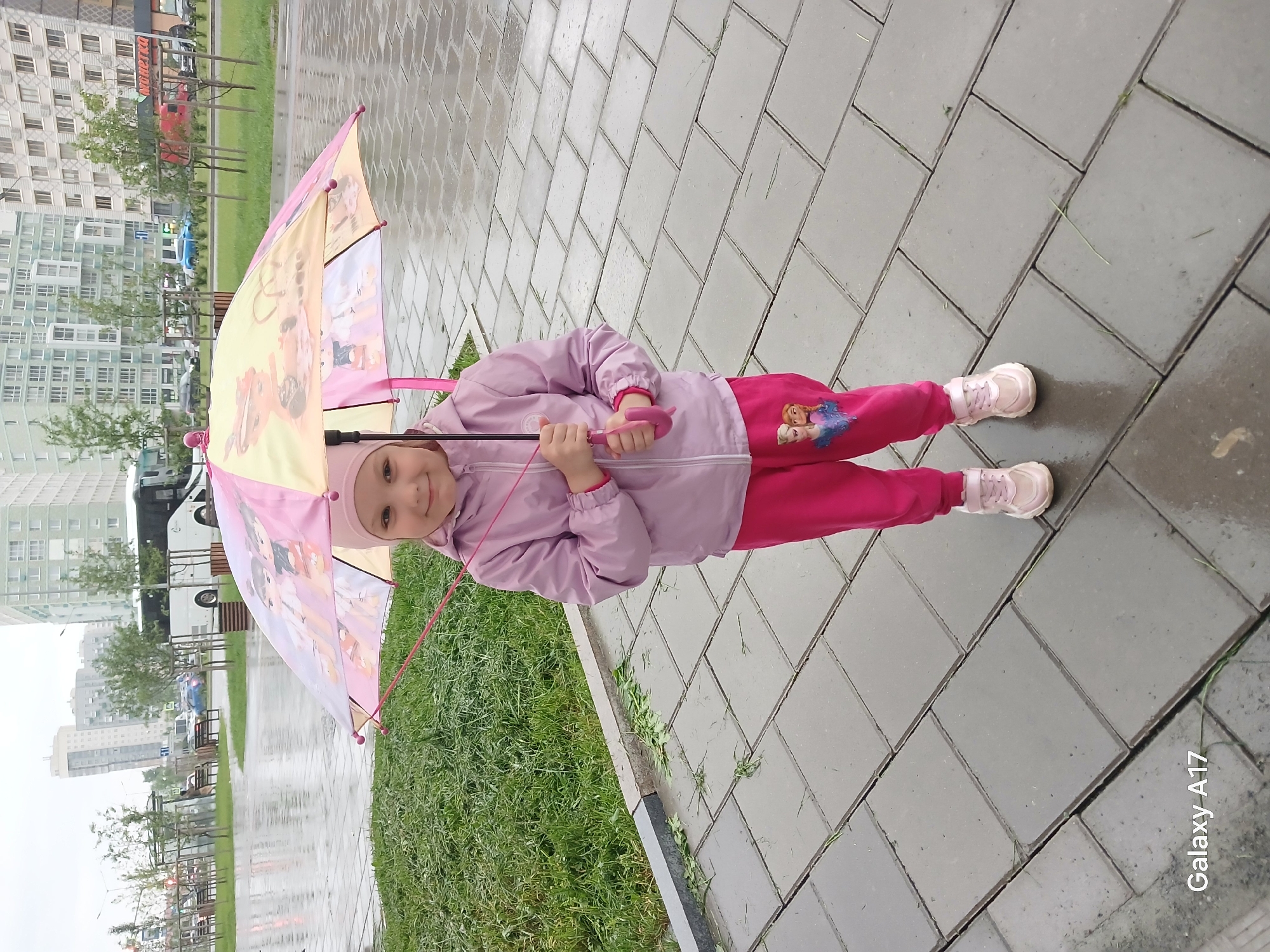 A young girl stands outdoors on a wet paved surface, holding an open, colorful umbrella. She is dressed in a light pink zippered jacket, bright pink sweatpants featuring a print of two female cartoon characters (likely Disney's Frozen princesses), light pink sneakers, and a matching pink hat or beanie. The child is smiling directly at the camera, conveying a sense of happiness and enjoyment. Her child-sized umbrella is primarily yellow and pink, adorned with various cartoon animal characters, possibly from My Little Pony.
The setting is an urban or suburban pedestrian area in Yekaterinburg, Russia. The ground beneath her is made of grey rectangular pavers, visibly wet, indicating recent rainfall. Patches of green grass line the pathway, and some green debris, possibly grass clippings, is scattered on the pavement. In the background, modern multi-story buildings rise under an overcast sky, suggesting daytime. Faintly visible cars and what appears to be a bus are in the distance, alongside trees and hints of street furniture like benches. A prominent red building in the far background clearly displays the name "Пятерочка" (Pyaterochka), identifying a well-known Russian supermarket chain. A "Galaxy A17" watermark is visible in the bottom right corner of the image.
