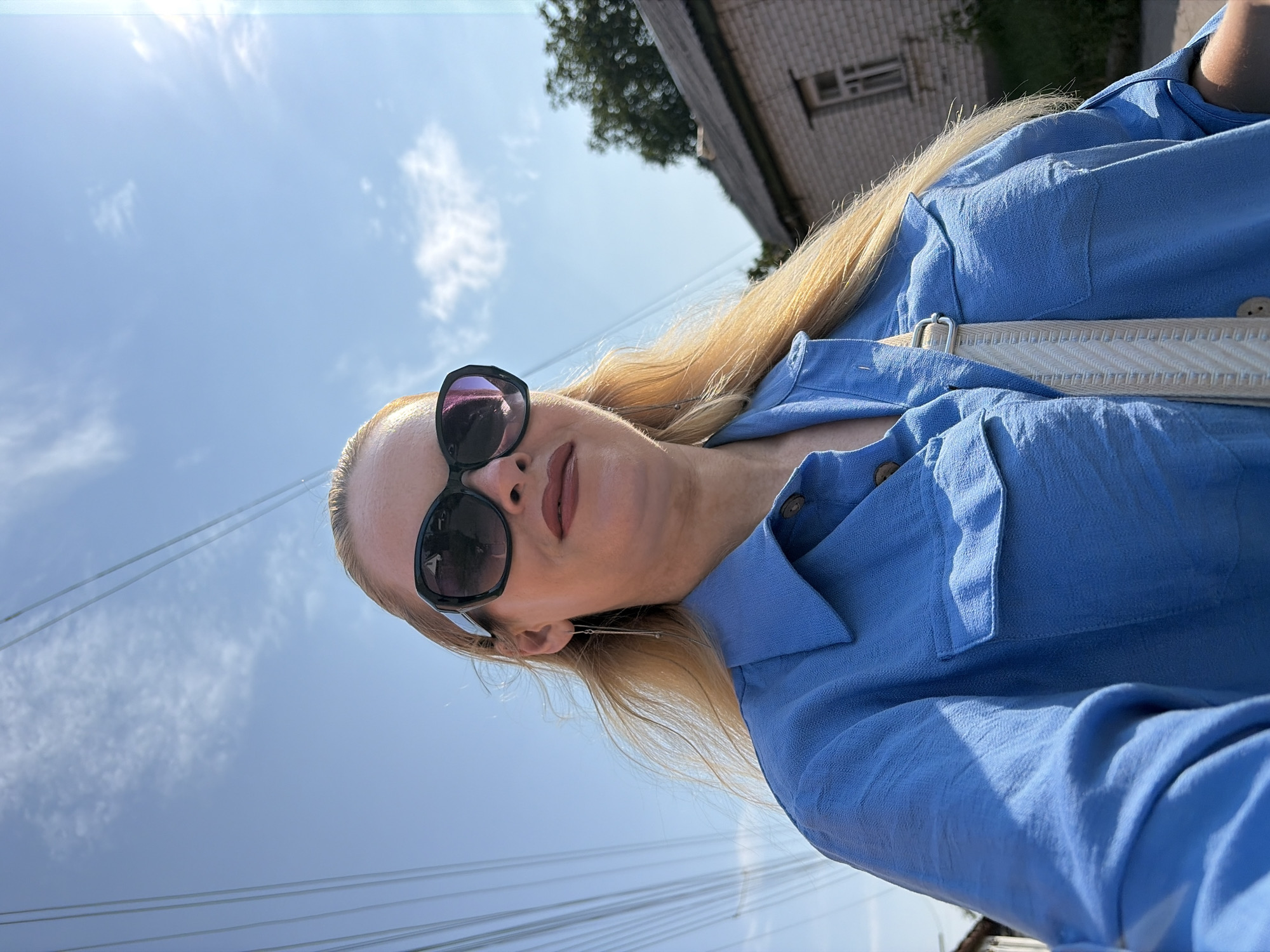 A blonde woman wearing large, dark sunglasses and a blue button-up shirt stands outside under a bright, partly cloudy sky. She has long, blonde hair that is slightly windswept. A white strap from a bag crosses diagonally over her chest. In the background, power lines are visible against the sky, and a simple brick building with a sloped roof can be seen. The overall setting appears to be a residential or rural area, likely on a sunny day.