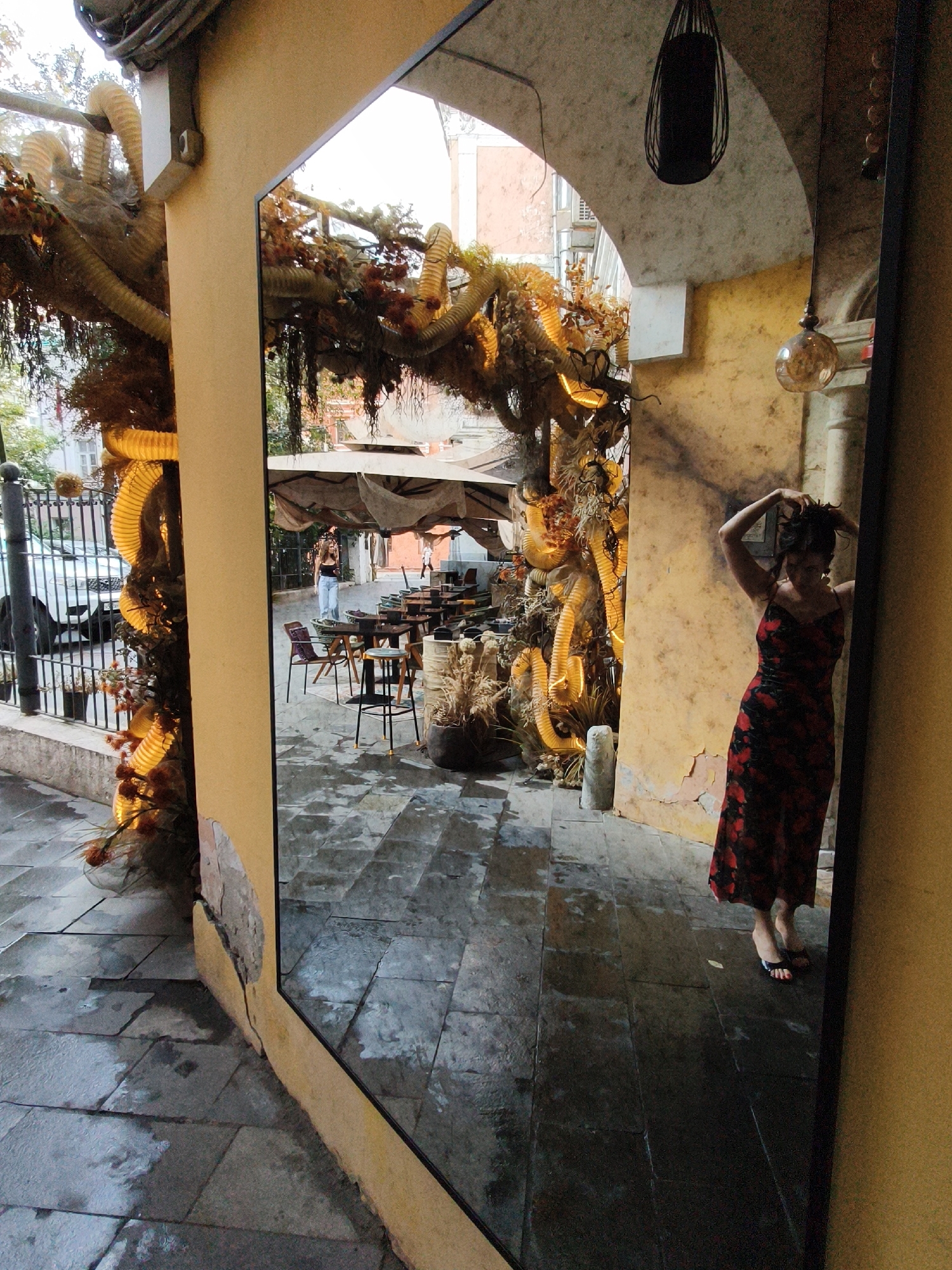 A full-body shot captures a woman in a long, red floral-print dress and black sandals standing next to a large mirror. She is posing with her hands in her dark hair, looking down and to her left. Her posture suggests she is aware of being photographed. The mirror reflects an outdoor seating area of a cafe, with tables, chairs, and patrons. The cafe is decorated with a large, elaborate, dried plant installation with yellow accordion-like elements and orange floral accents, which also appears in the foreground of the main image to the left of the mirror. The ground is paved with large, irregularly shaped grey stones, some of which are wet and reflective, indicating recent rain. The wall behind the woman has peeling yellow paint. The scene is lit by artificial lights and daylight filtering through. The mirror is framed by a beige wall to the left and a dark frame to the right. The image appears to be taken in an urban environment, possibly a courtyard or an alleyway with a cafe. The style of the building and the foliage suggests it could be in a Mediterranean or Southern European city. However, without additional information or context, pinpointing the exact location in Moscow, Russia is not possible.