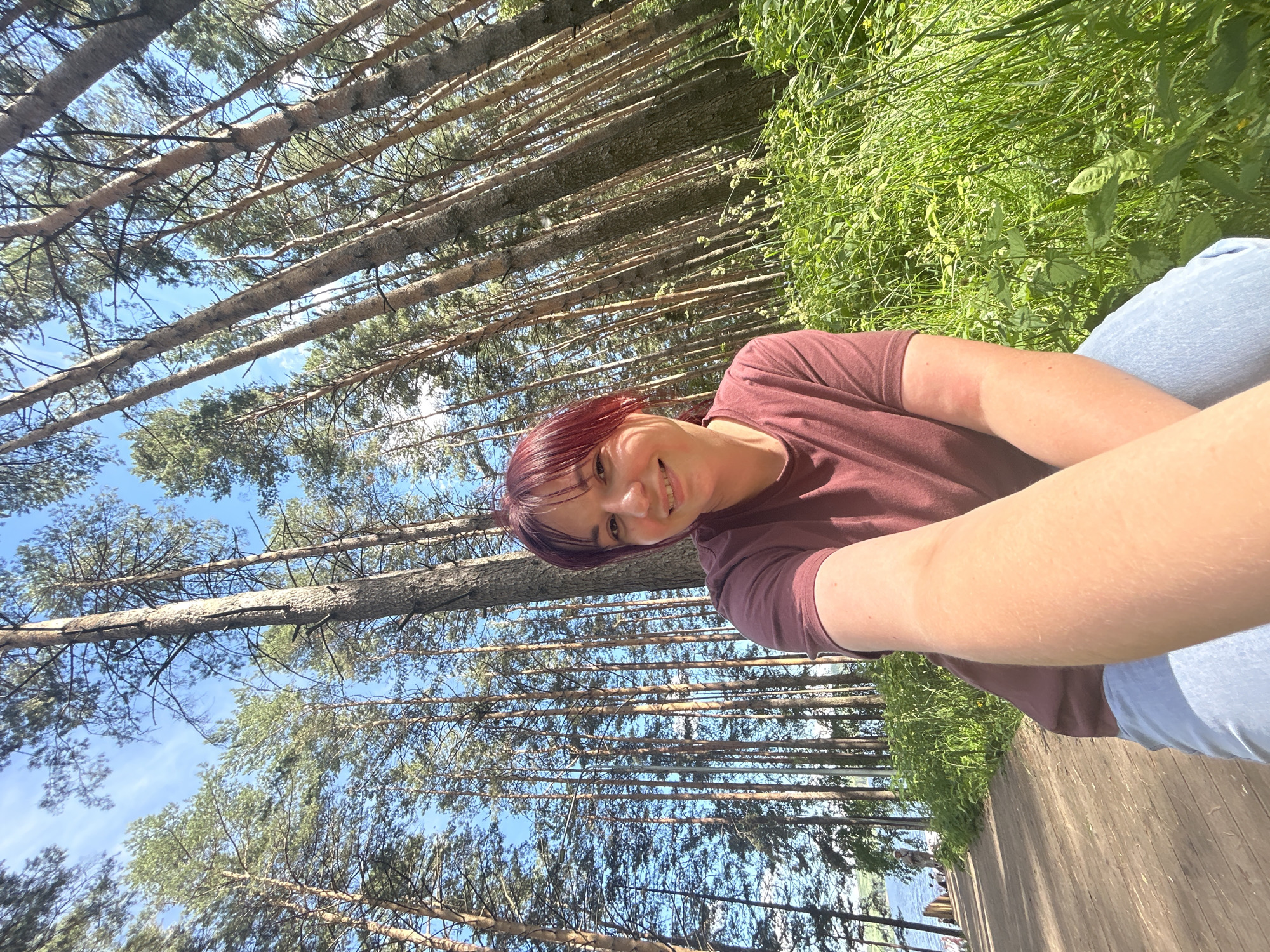 A person with short, reddish hair smiles at the camera in a selfie taken outdoors. They are wearing a maroon t-shirt and light blue jeans. The background consists of tall pine trees with clear blue sky visible between their branches. To the right of the person is lush green grass and foliage. In the lower portion of the image, a wooden boardwalk is visible, and beyond that, a body of water with a few people on the shore. The sunlight suggests it is daytime, and the clear sky indicates pleasant weather. The overall scene is natural and relaxed, possibly depicting a leisurely outing in a park or by a lake. There is no visible text in the image.