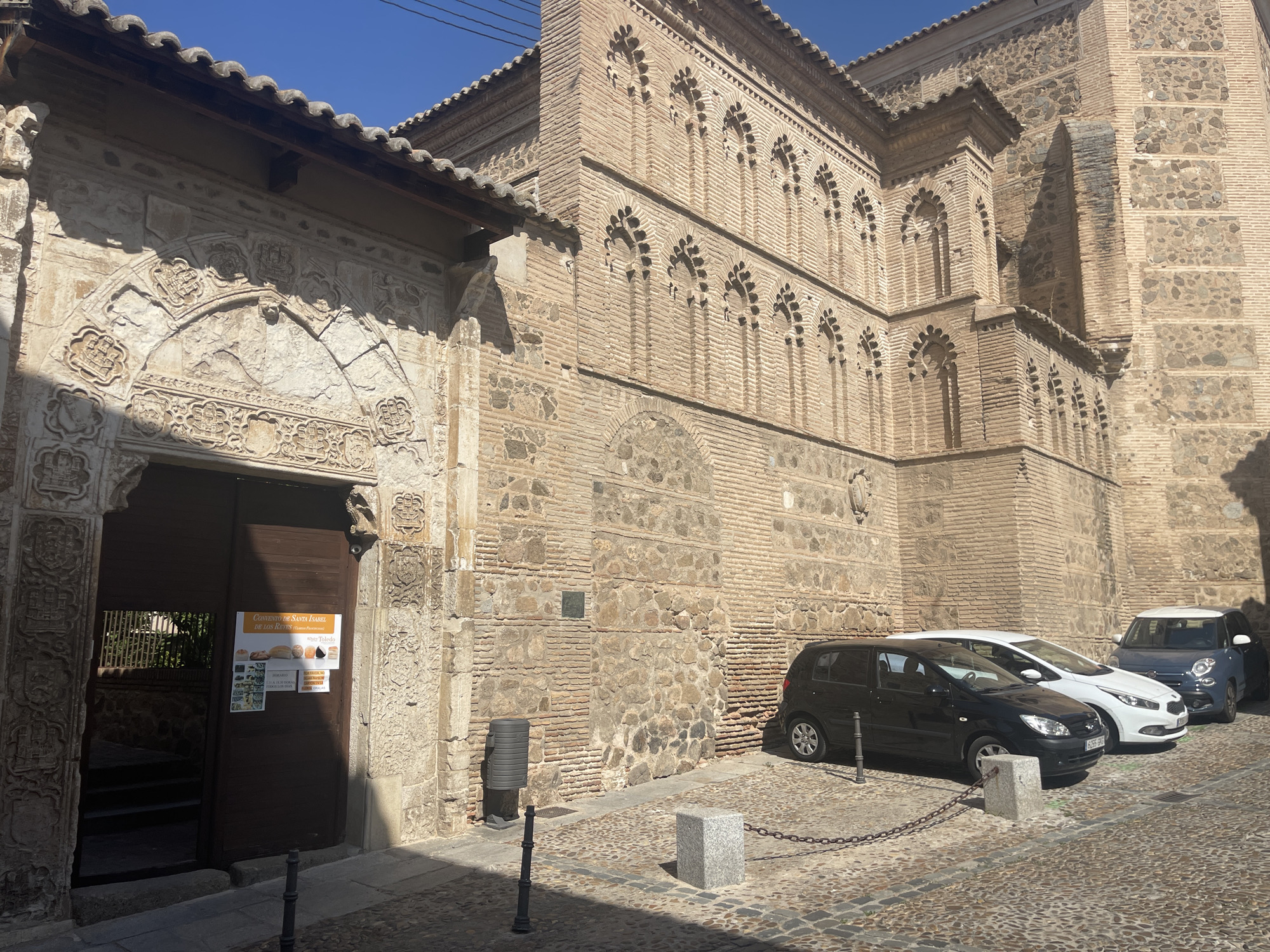The image depicts the exterior of the Convent of Santa Isabel de los Reyes in Toledo, Spain, on a sunny day. The architecture features a blend of stone and brick, with intricate carvings above the entrance. The entrance itself is a large, dark wooden door with a poster on it. To the right of the entrance, several cars are parked along a cobblestone street, separated by a chain barrier. The surroundings include more brick buildings with decorative arched windows. The sky is clear and blue.