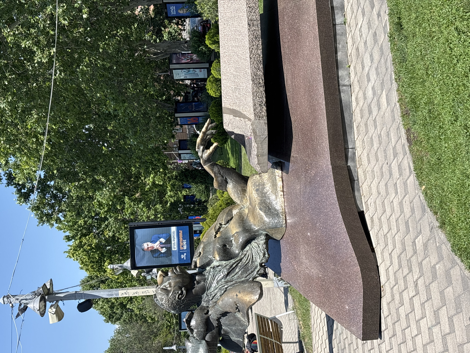 The image depicts a bronze sculpture of a man reclining on a stylized bench. The sculpture is situated in an outdoor urban environment in Yerevan, Armenia, likely a public park or square given the presence of trees, paved walkways, and grassy areas. In the background, there are posters or digital displays, one of which shows a portrait of a man in a suit and tie, with Armenian text below. The sculpture itself is abstract and fragmented, with flowing lines and an expressive pose. The man in the sculpture is leaning back, with his head tilted upwards and his left arm outstretched, appearing to gesture or point. The base of the sculpture is made of polished dark red granite, and the bench part is a rough-textured grey stone. A park bench with a person seated is visible to the left of the sculpture. The overall lighting suggests a sunny day.