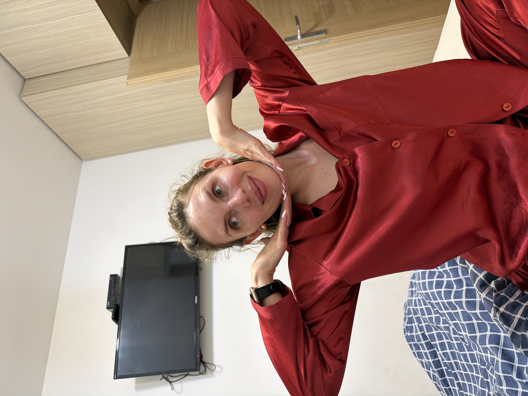 A fair-skinned woman with freckles is visible. She is wearing a maroon silky pajama set. She has her hands on her neck and chin, posing for the picture. She is wearing a smartwatch on her left wrist. Behind her is a mounted black television and a cabinet above the television, and the room has white walls. The blue and white checkered bed sheet is at the bottom right corner of the image. The location is assumed to be indoors, possibly a hotel room in Budva, Montenegro.