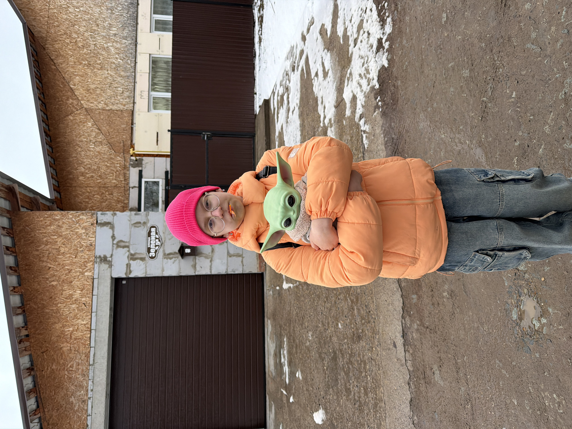 A girl is holding a Baby Yoda doll. The girl is wearing a pink hat, glasses, an orange jacket, and blue jeans. The location is in Chaikovsky, Russia. There is a garage door and a building in the background. There is snow and pavement on the ground.