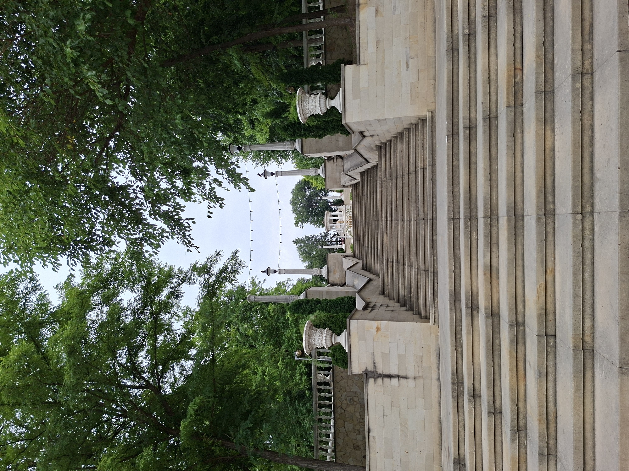 The image depicts a grand stone staircase leading up to a pavilion at the entrance of the Alley of Classics in Chișinău, Moldova. Lush green trees frame the scene, with the overcast sky suggesting a cloudy day. The staircase is flanked by decorative urns and classical columns. In the distance, the open-air pavilion is visible, a landmark of the city's cultural landscape. No people are present in the image.