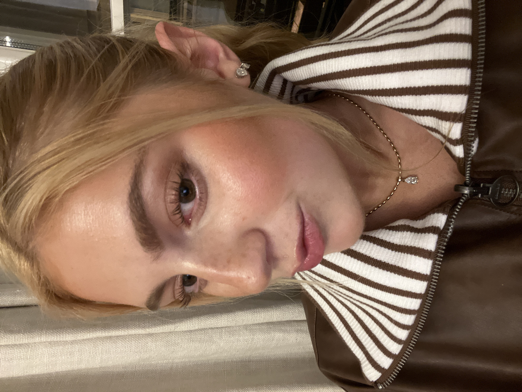 A close-up, tilted shot captures a blonde-haired woman with fair skin. She wears a brown and white striped garment under a brown leather jacket. A gold necklace with a pendant adorns her neck, and a stud earring is visible in her ear. The background features a white textured fabric. The image does not provide enough information to determine if the scene is in Paris, France.