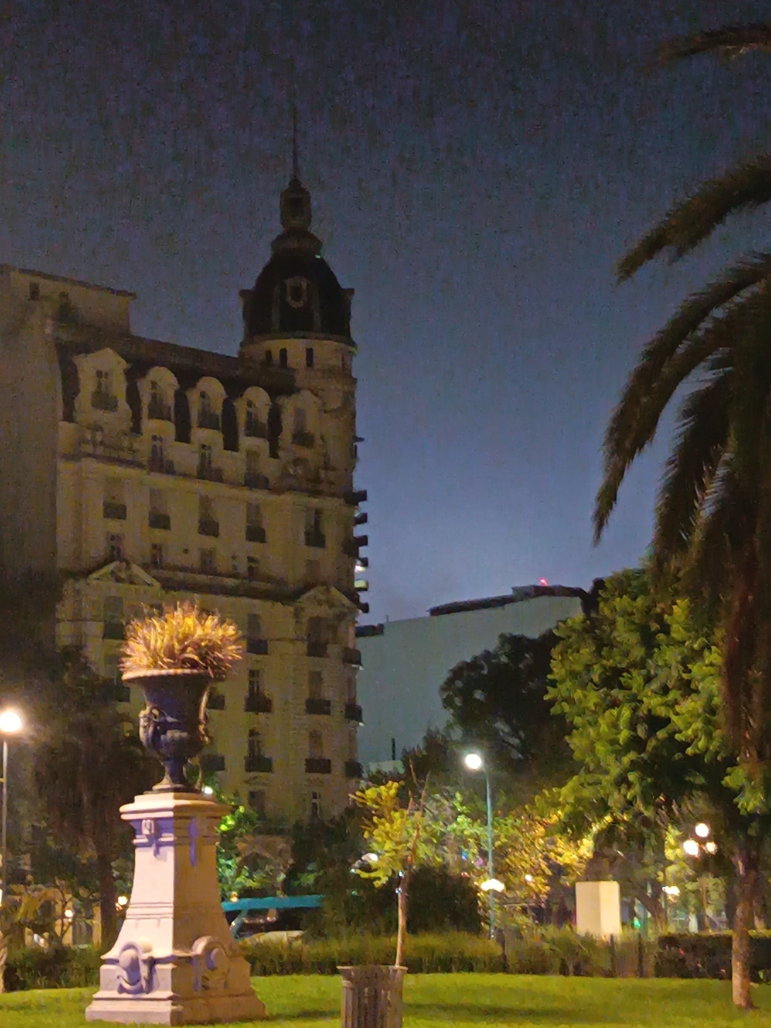 The image depicts a nighttime scene in what appears to be a park in Buenos Aires, Argentina. Prominently featured is a tall, ornate, European-style building with a tower. A large classical-style flowerpot sits on a pedestal in the foreground, illuminated by a light source. The park is filled with trees and greenery, and streetlights provide additional illumination. A palm tree is on the right side of the frame. Other buildings can be seen in the background behind the trees.
