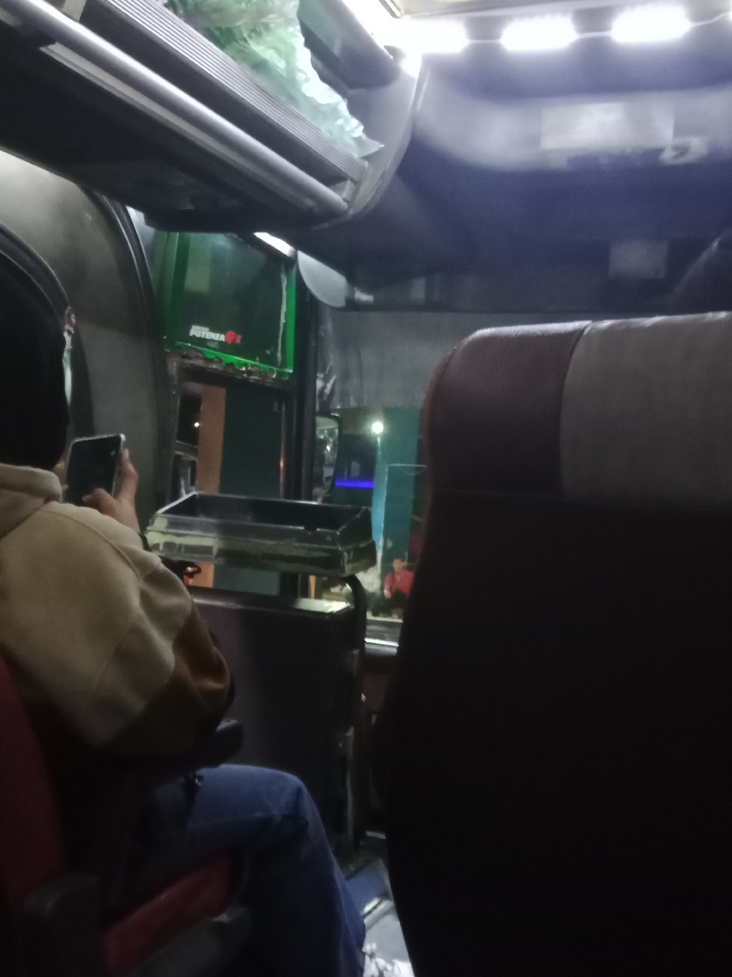 The image depicts the interior of a vehicle, possibly a bus, at night. In the foreground, a person wearing a tan and white hooded jacket and blue jeans is seated and holding a black smartphone in their right hand. The person is facing away from the camera, looking towards a transparent tray or container in front of them. To the right, a dark brown and tan seat is visible, partially obscuring the view of the person. In the background, there is overhead lighting and some green signage that appears to read "POTENZA" with a red logo. The overall lighting suggests it is dark outside, and the scene is illuminated by artificial lights within the vehicle. The specific location in the city of Surakarta, Indonesia, cannot be determined from the visual information provided in the image.