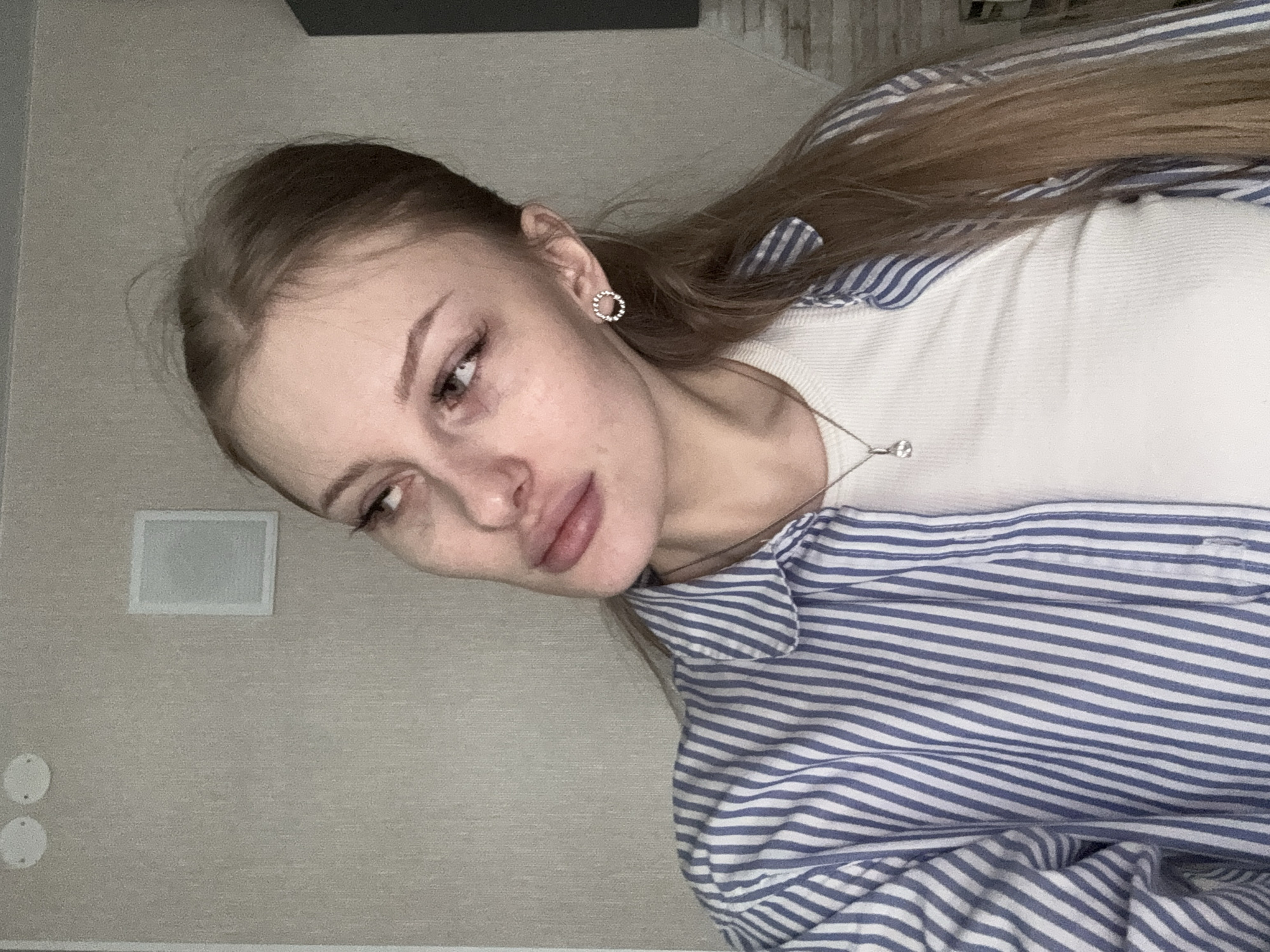 This is a close-up portrait of a young woman, likely a selfie, taken from a slightly elevated angle with her head tilted to her left. She has long, light brown hair that falls over her shoulder, and she wears a subtle circular stud earring adorned with small gems in her right ear. Her expression is calm and natural, with her eyes gazing downwards and to her left, and her lips slightly parted. She has well-defined eyebrows and appears to be wearing minimal or no makeup.
Her attire consists of a white, subtly ribbed long-sleeved top layered under a blue and white striped button-up shirt. A delicate silver necklace with a small, clear pendant hangs around her neck.
The background features a light, textured wall, possibly in a shade of beige or light grey. On the wall, there is a rectangular white object, which could be a speaker or a wall-mounted fixture, and two smaller circular white covers below it. A darker, possibly wooden or tiled surface is visible in the upper part of the frame, suggesting an indoor setting. The lighting is soft, indicating an indoor environment. There is no visible text in the image. The location context for this image is Perm, Russia.
