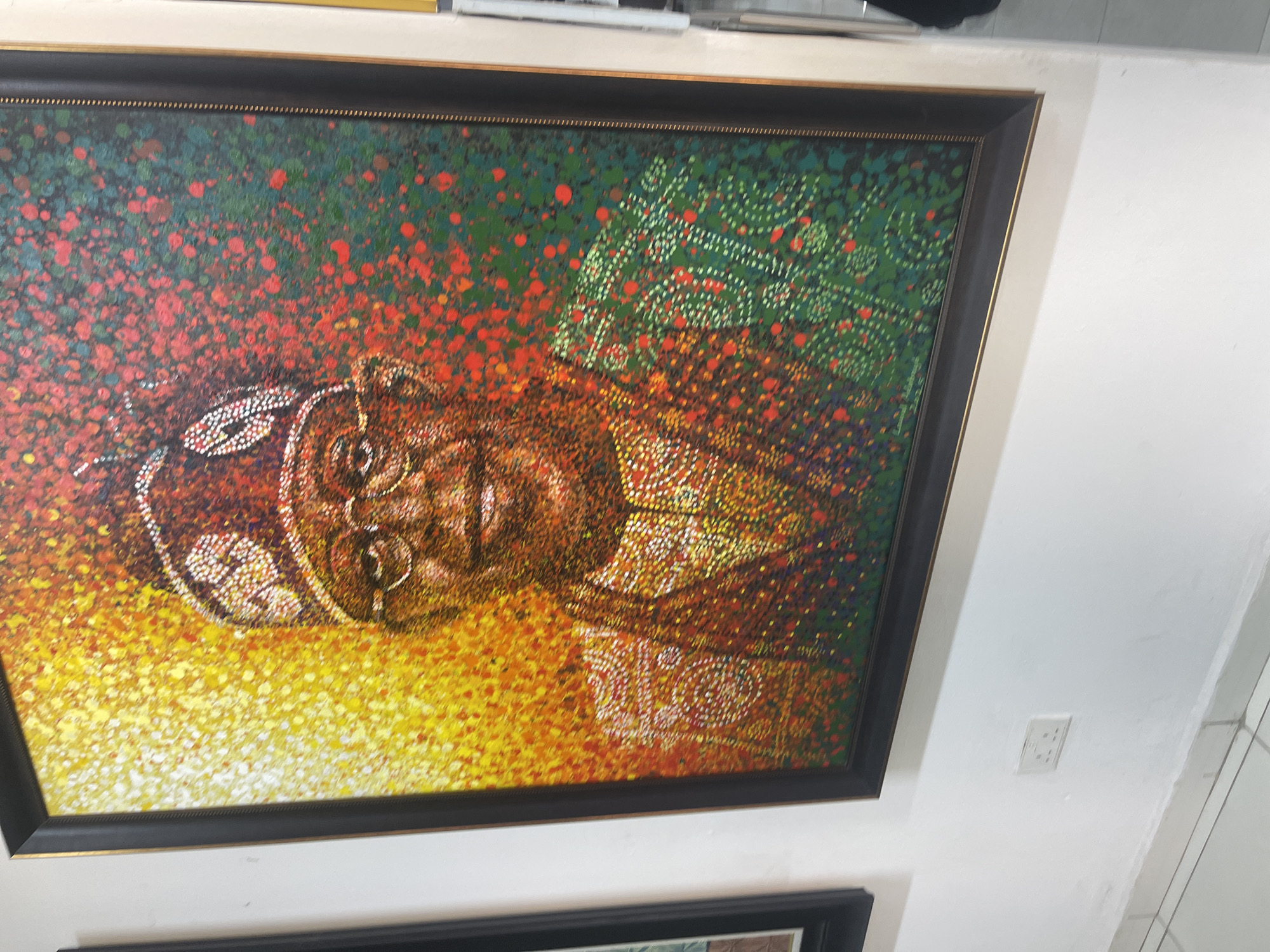 This media file showcases a large, vibrant pointillistic painting displayed on a plain white wall, likely in an indoor setting in Abuja, Nigeria. The artwork depicts a portrait of an individual, presumed to be a man of African descent, shown from the chest up. He is adorned in traditional attire, featuring a patterned cap or headwrap and an intricately designed garment. The subject is wearing glasses and bears a gentle smile.
The painting's distinctive style utilizes numerous small, distinct dots of color to form the image. The background transitions from vivid reds and oranges in the upper left, suggesting warmth, to bright yellows at the bottom. The upper right quadrant features a dark green background with elaborate patterns composed of lighter green and white dots. The individual's face and clothing are rendered with a blend of warm tones and white dots, emphasizing the detailed patterns on his attire and headwear.
The artwork is set within a dark frame, possibly black, edged with a slender gold inner border. A portion of another framed artwork can be seen below it. To the right of the main painting, a white electrical outlet is mounted on the wall. In the lower right corner of the painting, a signature or inscription made of white dots is visible, appearing to read "DIGO" or "DIGBO" followed by other less decipherable marks. The painting conveys a sense of cultural richness and artistic vibrancy.