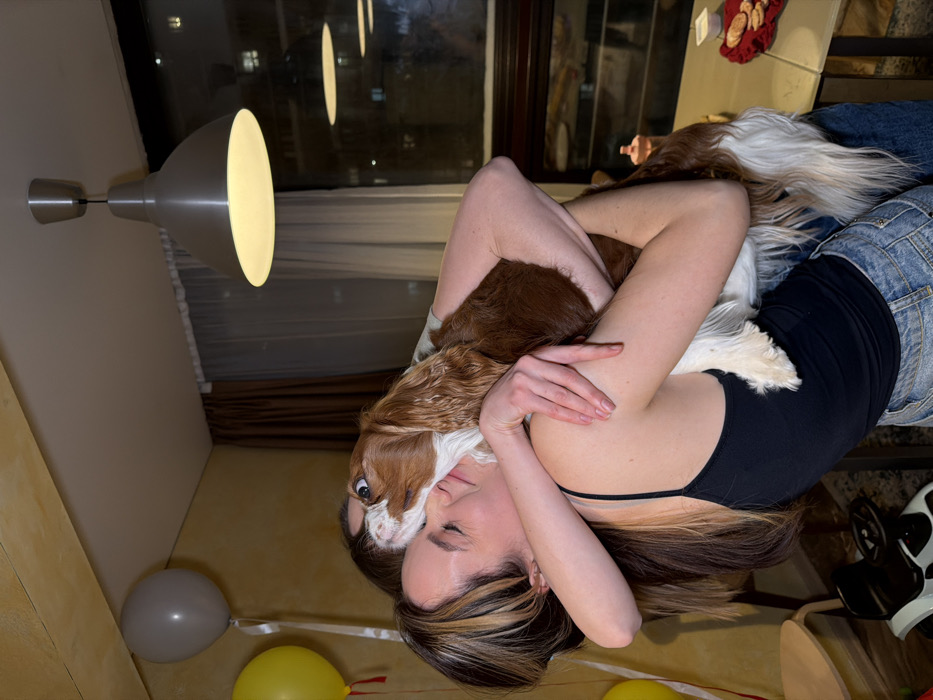 A woman embraces a Cavalier King Charles Spaniel. She wears a black tank top and denim shorts. The dog rests on her chest, its head near her face. In the background, a window reflects interior lighting, and a table with a tray of pastries is visible. Below, balloons are scattered on the floor. A modern wall lamp illuminates the scene from the side. A toy is positioned on the right.