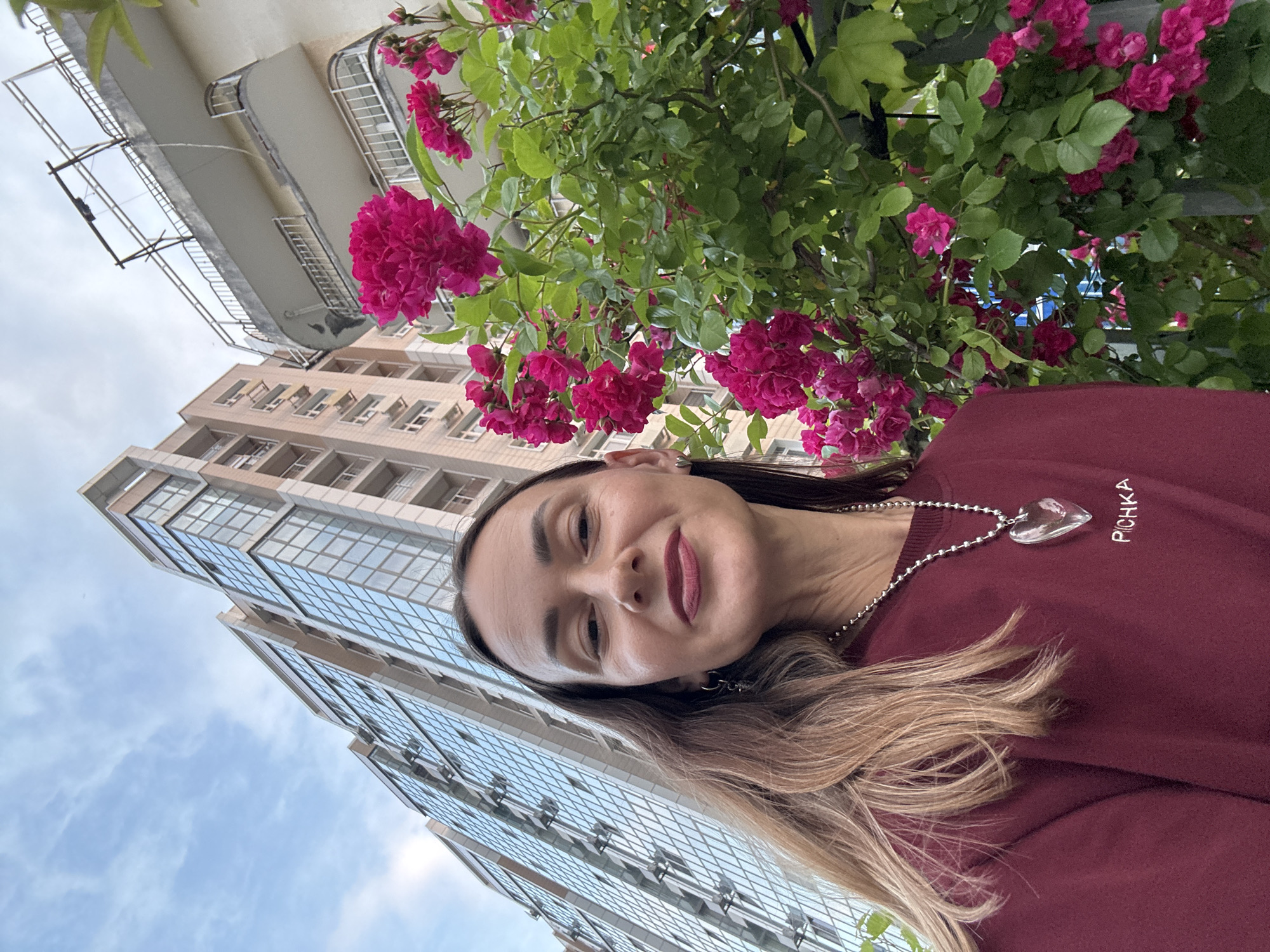 A low-angle, outdoor, eye-level, medium-shot selfie shows a woman with long, brown hair and a maroon sweatshirt standing in front of a rose bush with pink flowers and a modern building in the background. The woman is looking directly at the camera with a slight smile. She is wearing a silver necklace with a clear, heart-shaped pendant and a silver hoop earring in her left ear. The rose bush is in full bloom, with many clusters of vibrant pink flowers and green leaves. Behind the rose bush, a tall, modern building with a glass facade rises towards the sky. To the right of the modern building, an older, cream-colored building with balconies is visible. The sky is partly cloudy with a pale blue hue. The scene appears to be in a city environment, possibly a residential area. The image was captured in Sochi, Russia.