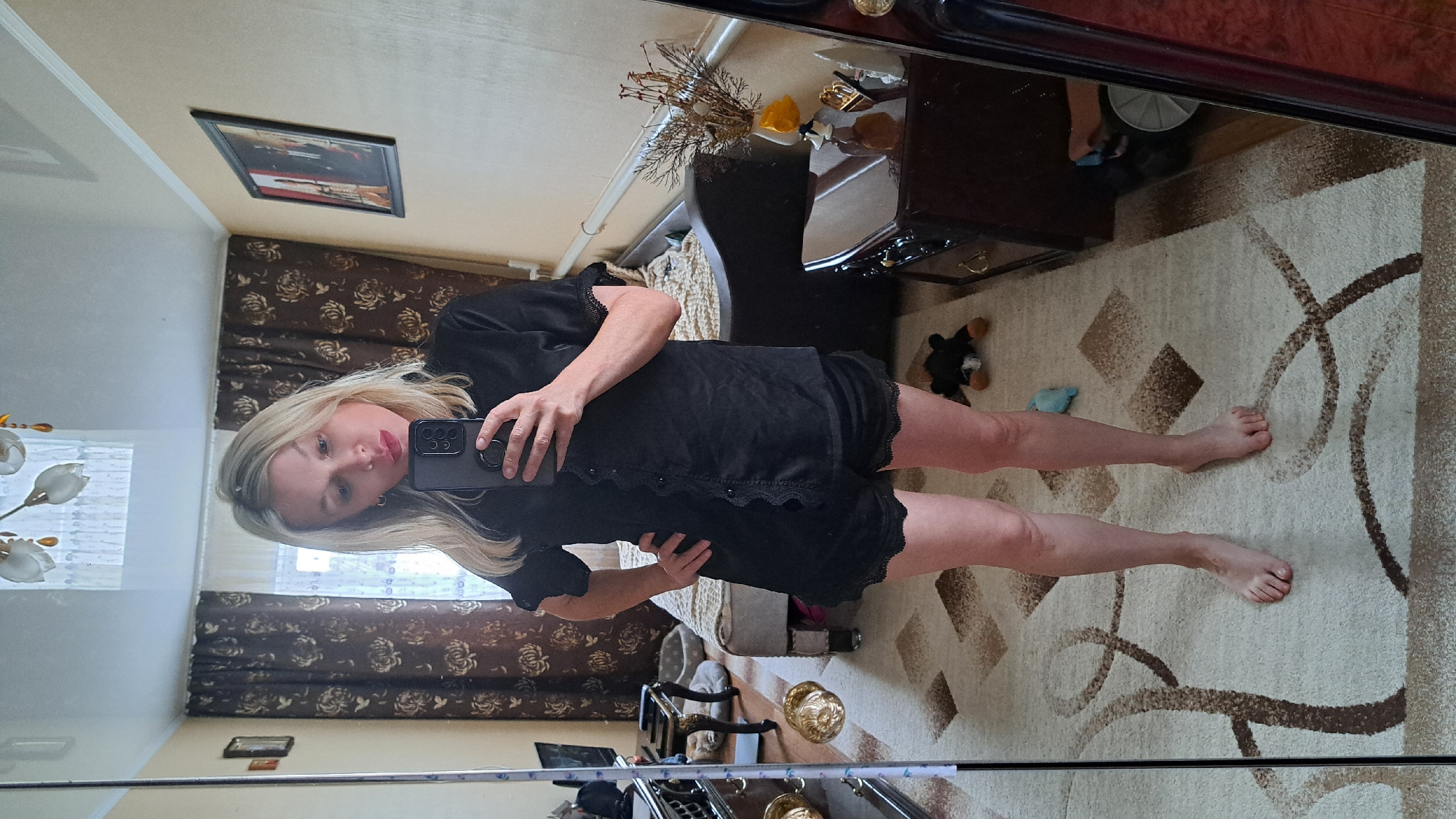 A blonde woman stands in a bedroom, taking a selfie in a mirror. She is wearing a black pajama set with lace trim. The room has a bed with a patterned comforter, a dresser with decorative items, curtains, and a rug on the floor. A framed picture is on the wall.