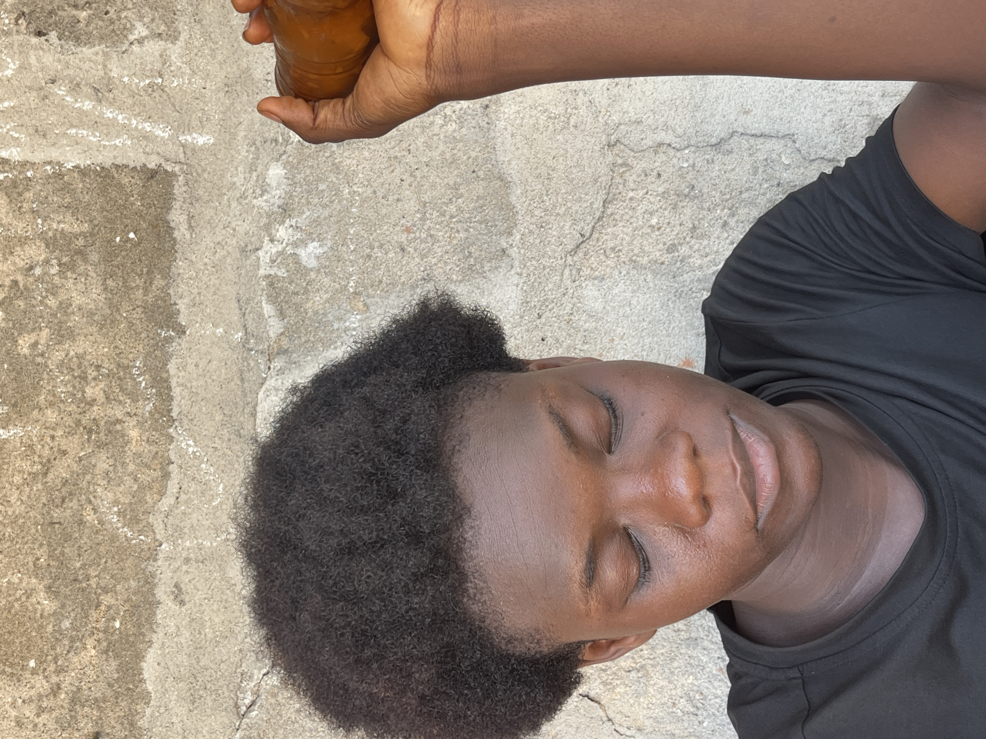 A medium shot, taken from above, depicts a person with dark skin and short, curly hair lying on a concrete surface. The person’s eyes are closed. They wear a black t-shirt. At the top of the frame, the person’s hand holds a brown object. The location, Badagry, Nigeria, is inferred. No action or interaction is occurring.