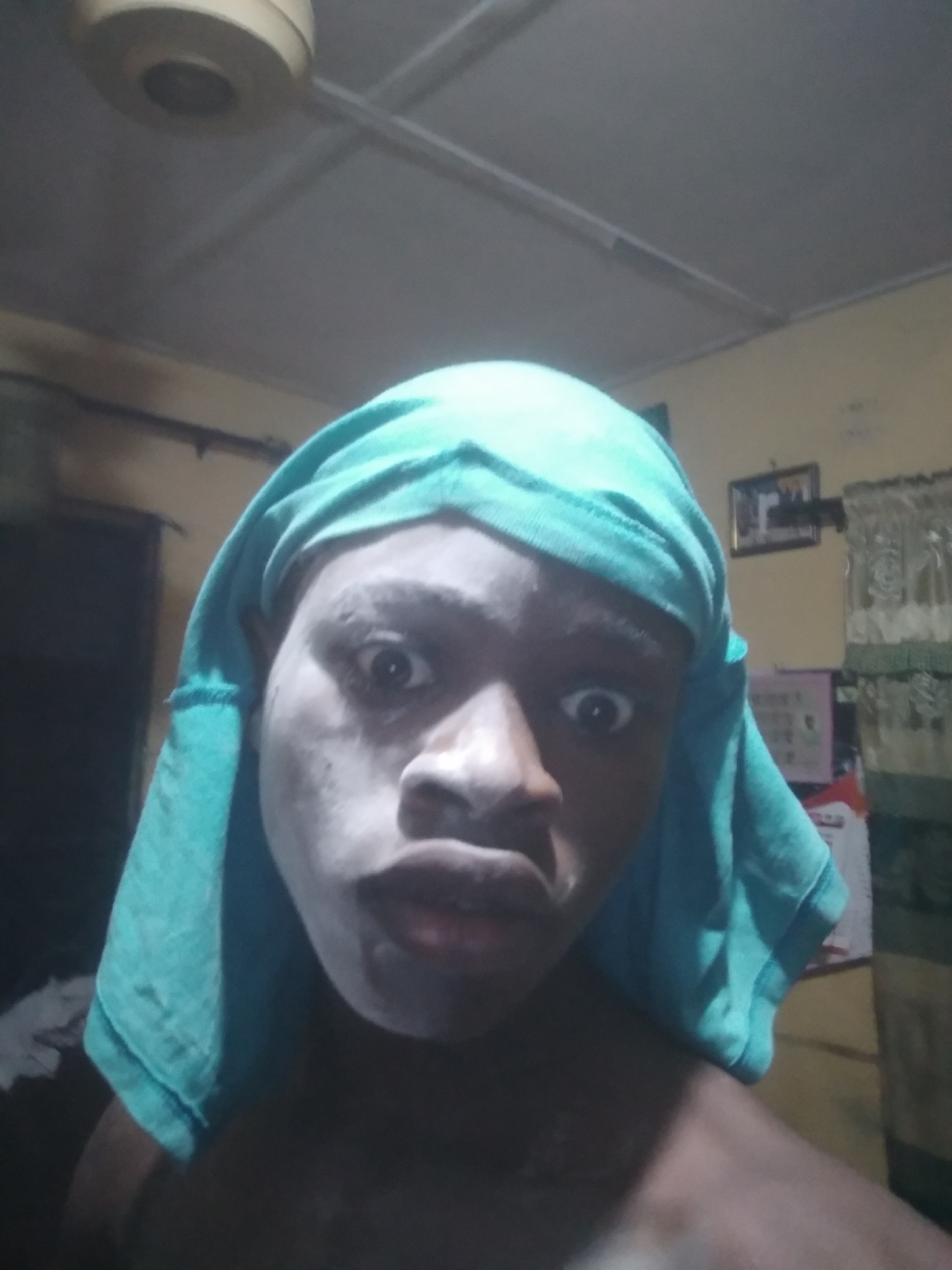 A dark-skinned individual is captured in a close-up, low-angle selfie within an indoor setting. The subject's face appears powdered with a white substance, and a turquoise fabric covers their head, draping down the sides. The person's eyes are wide and appear startled. The background shows a room with a ceiling fan, dark curtains, and a small framed picture. The location could be a room in a house in Shangisha, Nigeria.
