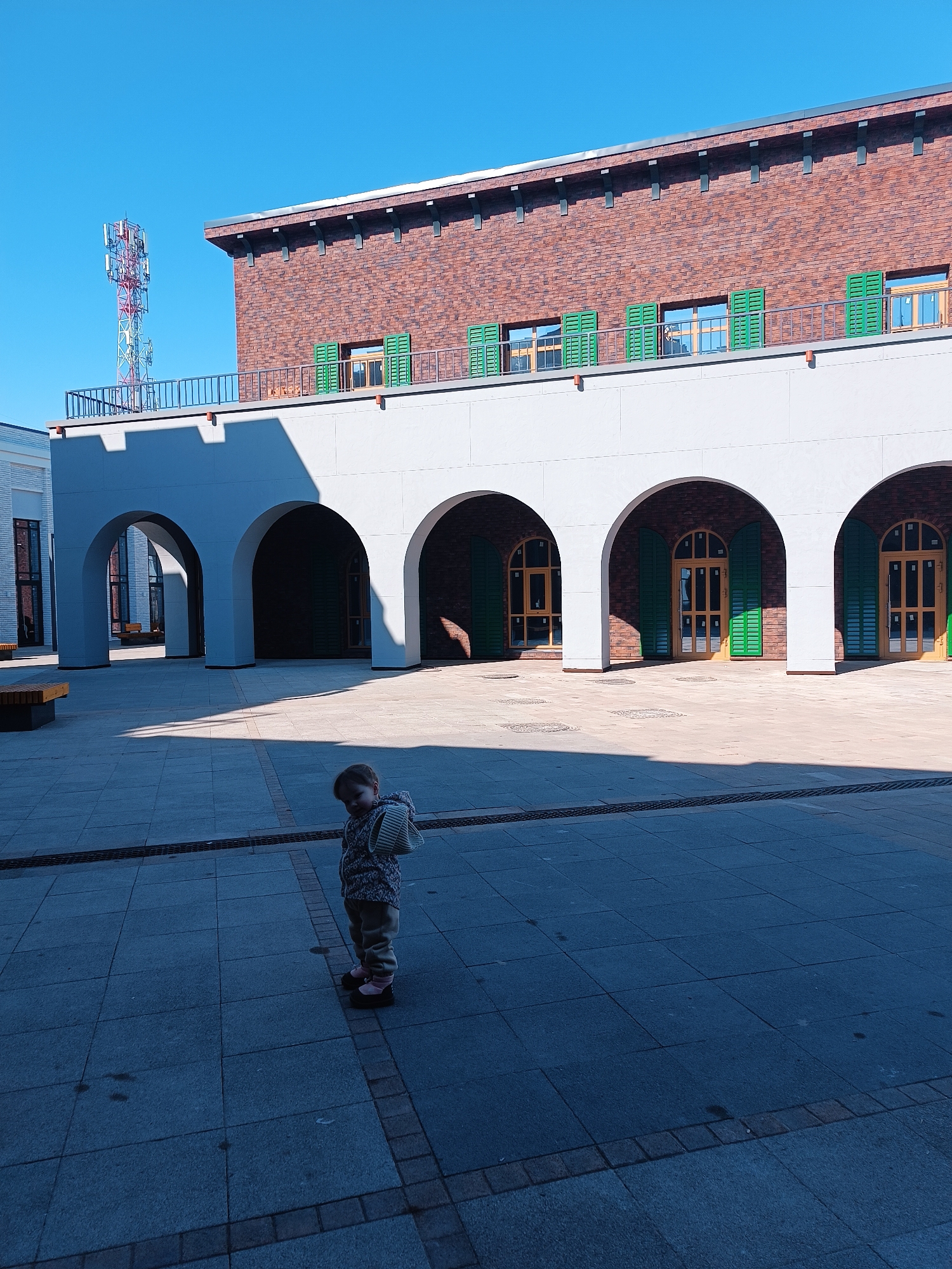 A low-angle, daytime shot shows a young child standing in an open plaza in Izhevsk, Russia. The plaza is paved with large, square tiles. The child wears a patterned sweater, pants, and dark shoes with pink socks. A small bag hangs off their shoulder. The child looks to the left side of the frame.
Behind the child is an architectural structure consisting of a light gray colonnade of arched openings with wooden doors featuring green shutters. Above this colonnade is a red brick building with a flat roof, black trim, and a metal railing. A communication tower stands on the left side of the frame behind the building. The sky is clear and blue. Shadows stretch across the plaza from the colonnade.