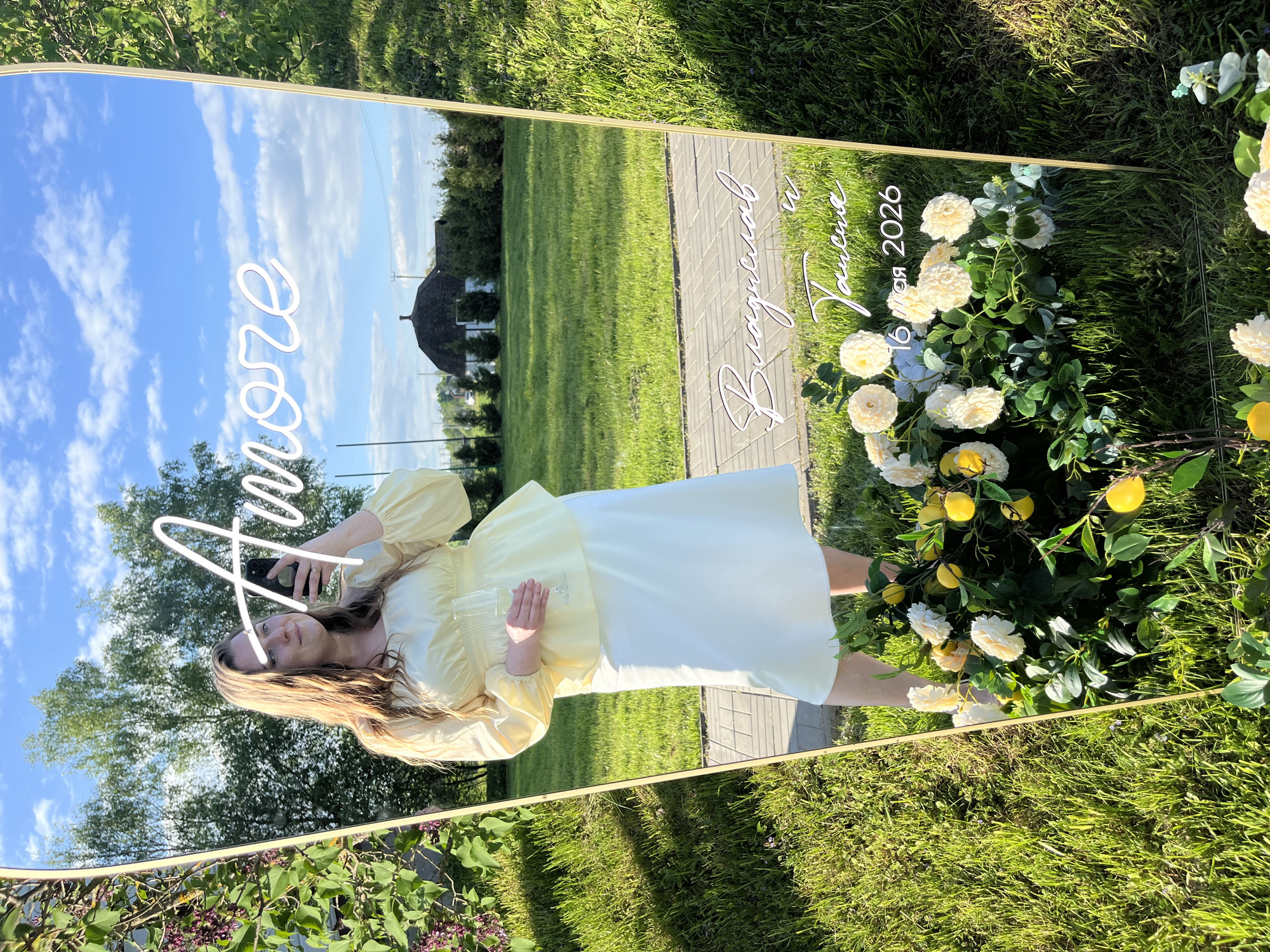 A woman is reflected in a large, tilted mirror placed outdoors on a grassy area, possibly in the city of Алопово, Russia. The mirror features decorative elements, including white floral arrangements and lemons near its base, and white, cursive writing across the top, including the word "Amore," and what appears to be a date written in Russian. The woman is holding a smartphone, suggesting she is taking a selfie. Her reflection shows she is wearing a yellow top and white skirt. In the background, the reflection in the mirror captures a clear sky with clouds, a tree, a section of lawn, and a building. The mirror has a gold-colored frame. The area around the base of the mirror features additional greenery and purple flowers.