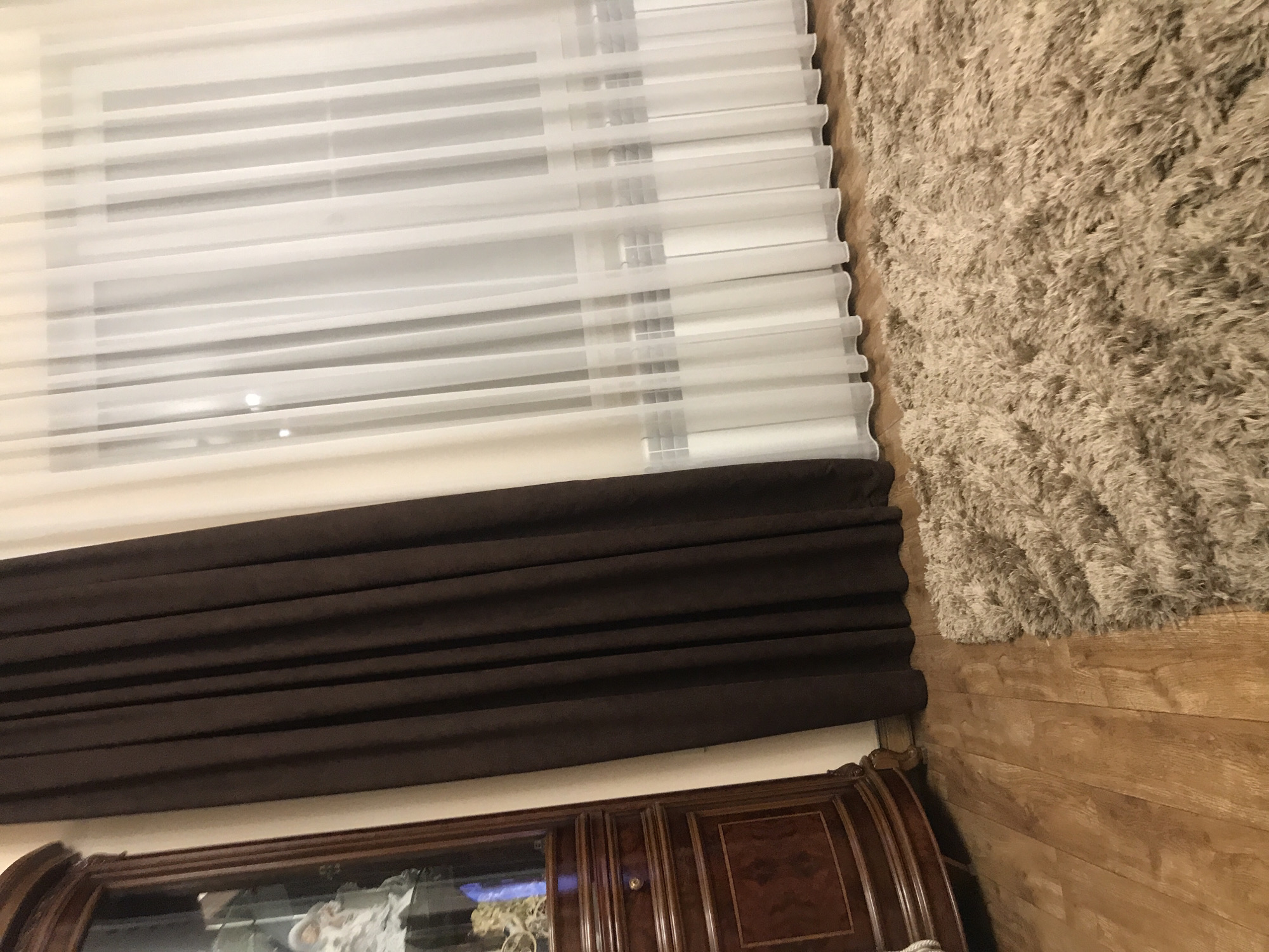 The image shows a room interior. A window covered by white blinds and a brown curtain is visible in the upper left. Below the curtain is a wooden cabinet with glass doors. To the right of the window is a tan shag rug on a hardwood floor. There is no evidence of the location being in Yerevan, Armenia. There are no people visible.