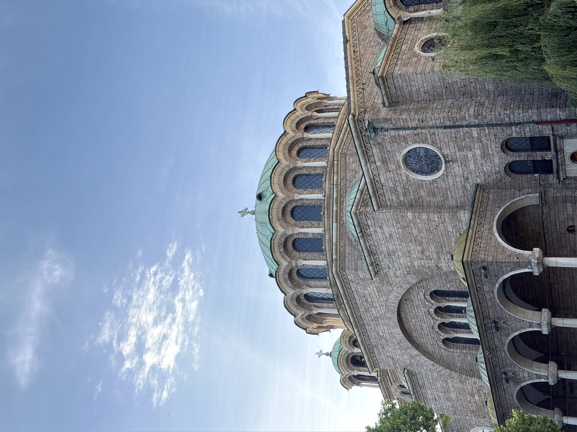 The image depicts the exterior of the St. Alexander Nevsky Cathedral in Sofia, Bulgaria, viewed from a low angle. The architectural details of the cathedral are prominently displayed, including the domes with their distinctive green patina, arched windows, and stone facade. The sky is light blue with wispy clouds. Trees are visible on the right and bottom edges of the frame, partially obscuring the building. No people are visible in the image.