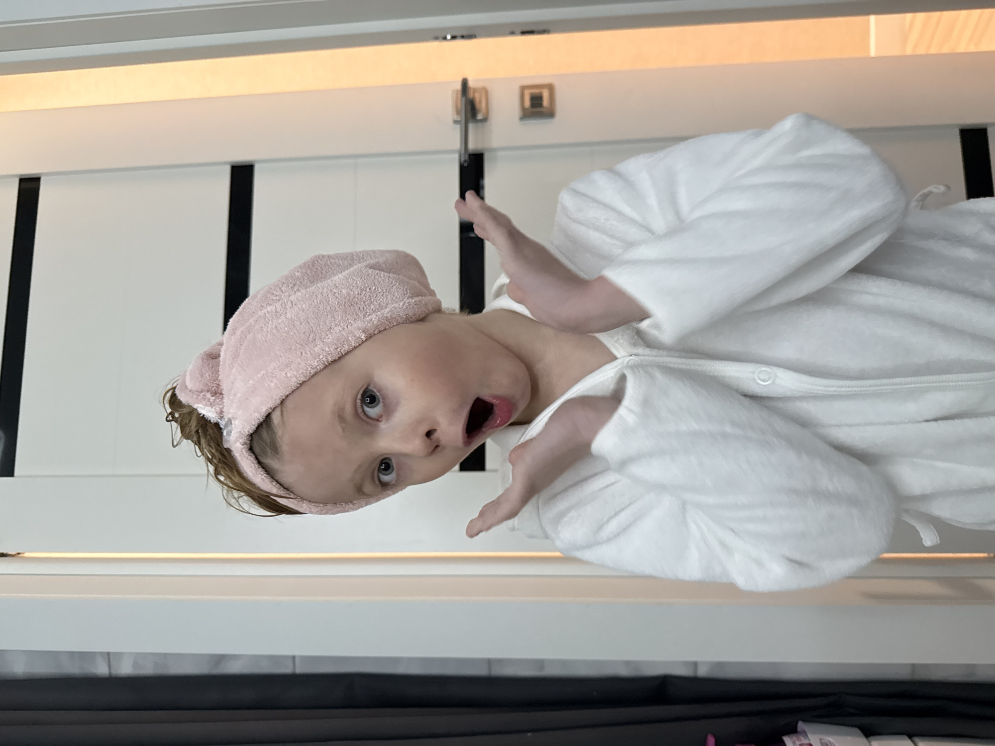 A person, presumably a child, is wearing a pink towel on their head and a white robe. They appear to be in a bathroom setting with white cabinets or paneling visible in the background. The person's expression is one of surprise or shock, with their mouth open and hands raised. The image does not contain location information to determine if it was taken in Izhevsk, Russia.
