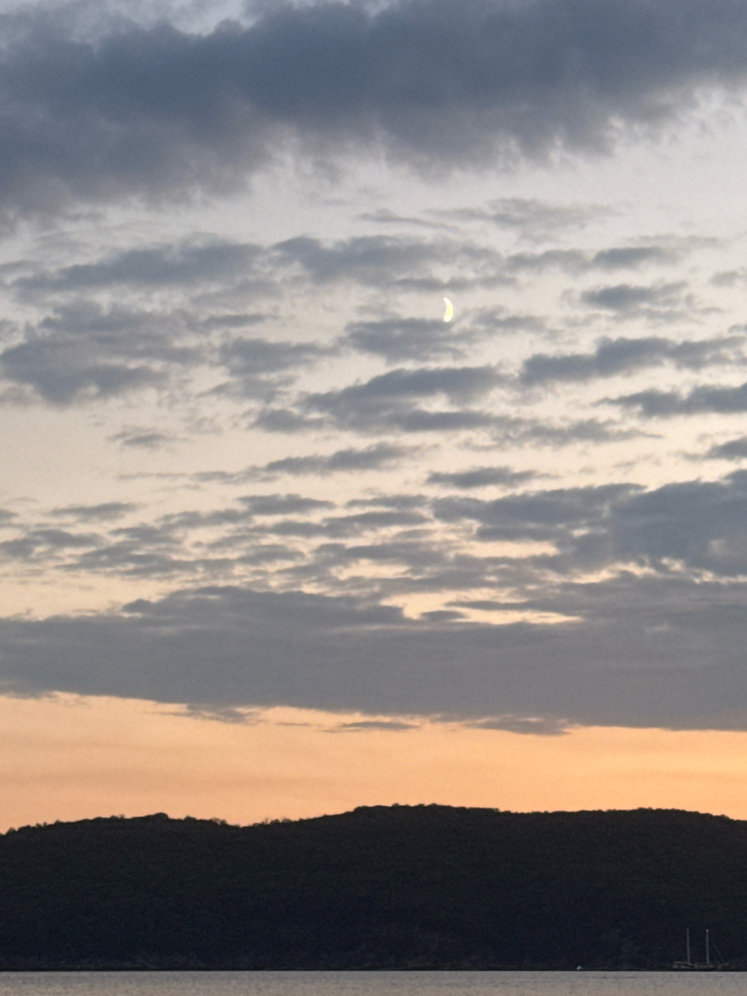 The image depicts a dusky sky with a crescent moon visible amongst scattered clouds. Below the sky is a dark, silhouetted treeline of a hill or mountain range. In the foreground, a body of water reflects the subdued light. There are no people or significant objects evident in the scene. The location is identified as Boreti, Montenegro.