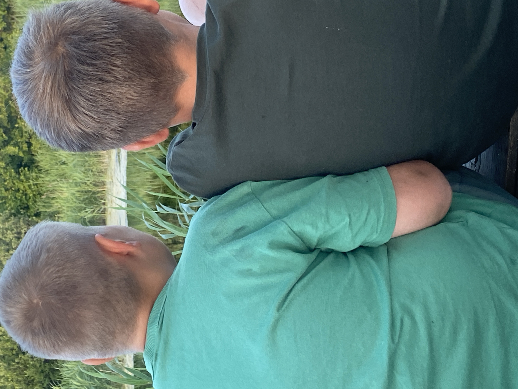 The image shows the backs of two young boys from the shoulders up, sitting side-by-side outdoors, facing away from the camera. The boy on the left wears a bright teal t-shirt, and the boy on the right wears a dark olive green t-shirt. Both boys have short, light brown hair. They are positioned in front of a natural backdrop of tall green reeds and foliage, with a body of water visible between the reeds. The scene suggests an outdoor setting, likely near a lake or river, in a rural or natural area. There is no indication of the specific location being within the city of Афанасьево, Russia.