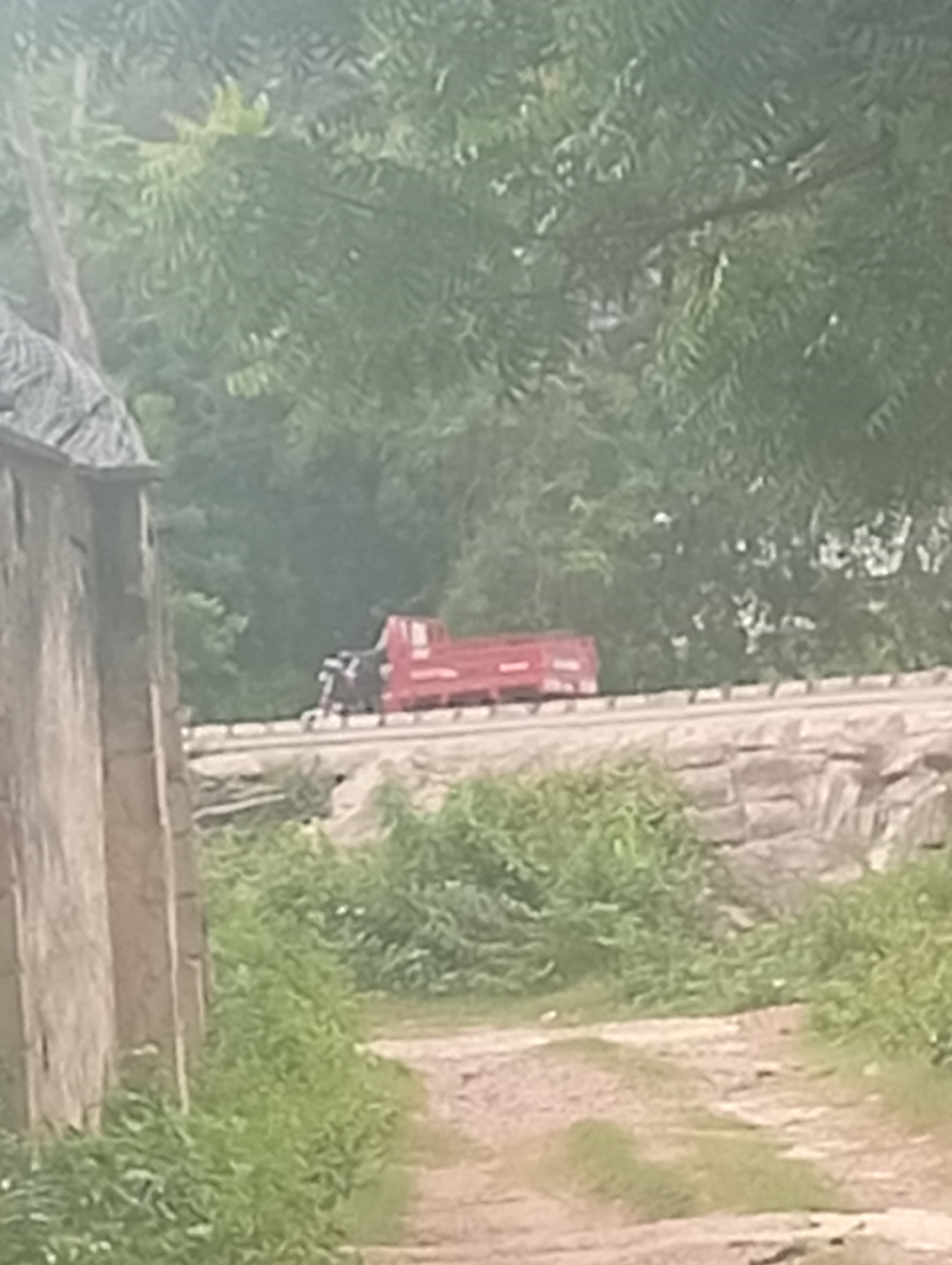 An outdoor, medium shot captures a red train car on a track, with trees and vegetation in the background. The train car is positioned horizontally in the frame, in the middle distance. The foreground features a dirt path with green grass and foliage on either side. To the left, a portion of a wooden structure, possibly a building or fence, is visible. The lighting suggests it is daytime, with diffused light, possibly indicating an overcast sky or a shaded area. The overall scene is rural or semi-rural.