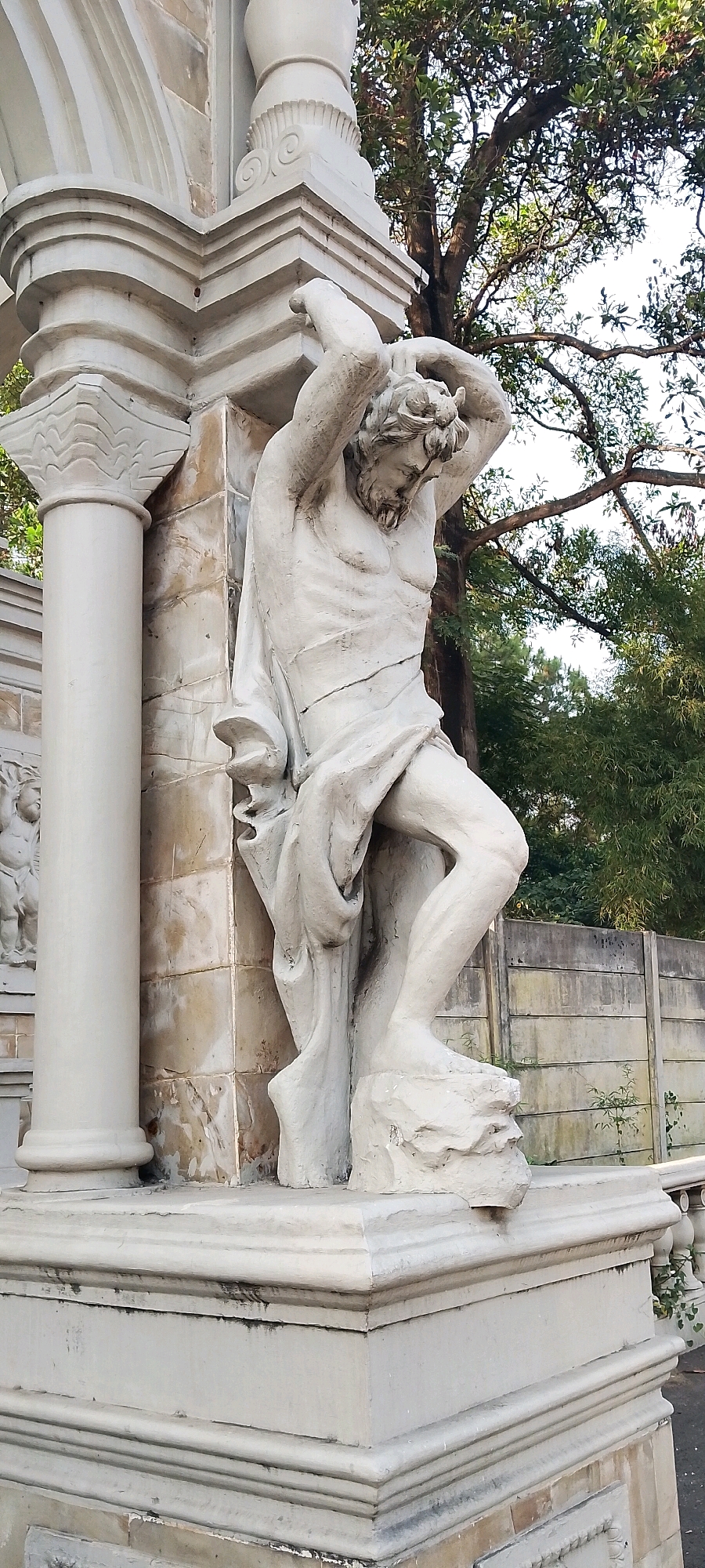 The image captures a close-up view of a weathered, light-colored stone statue depicting a muscular male figure, likely a titan or mythological character, with his arms raised and hands clasped behind his head, as if bearing a heavy load. The statue is positioned outdoors, next to a large, fluted column, and appears to be part of an architectural structure. The background consists of lush green trees and a textured, gray concrete wall. The lighting suggests it is daytime. The statue is in a state of disrepair, with visible cracks and chipped surfaces. The overall scene is one of an outdoor sculpture, possibly in a park or garden, with signs of age and exposure to the elements. The provided contextual information places this scene in the city of Gunung Putri, Indonesia.