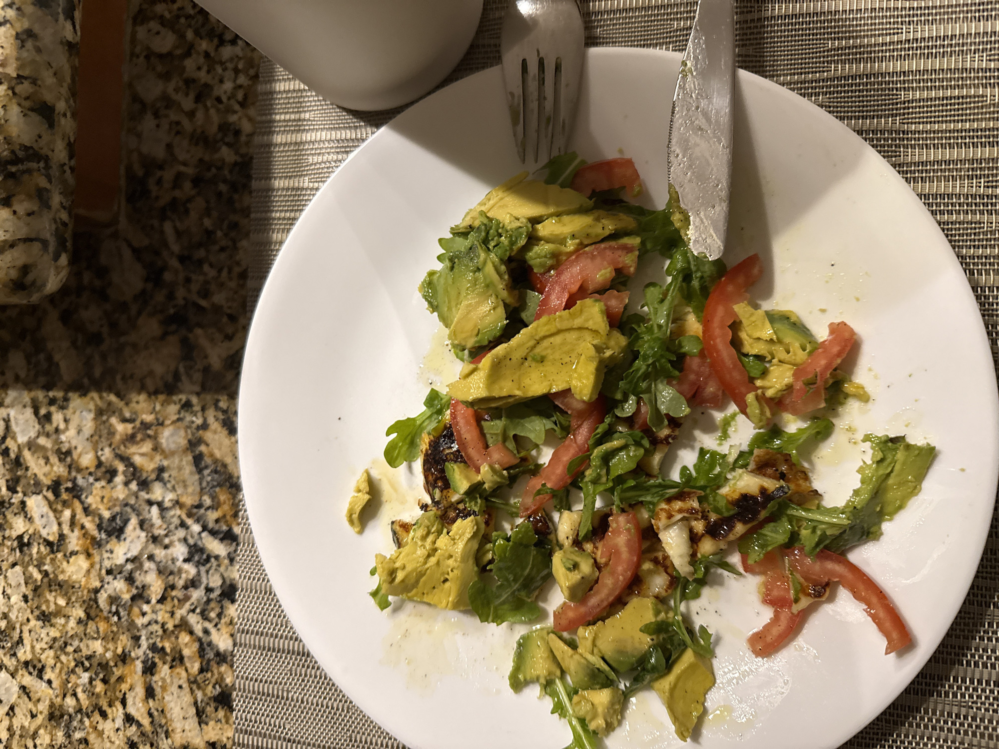 A partially eaten salad sits on a white plate, accompanied by a fork and knife. The salad consists of sliced avocado, tomato, and leafy greens. The plate rests on a textured surface with a light-colored container partially visible in the upper left corner. The presence of a knife and fork suggests that someone was eating the salad. The location of San Bernardino Tlaxcalancingo, Mexico is not discernible from the visual data.
