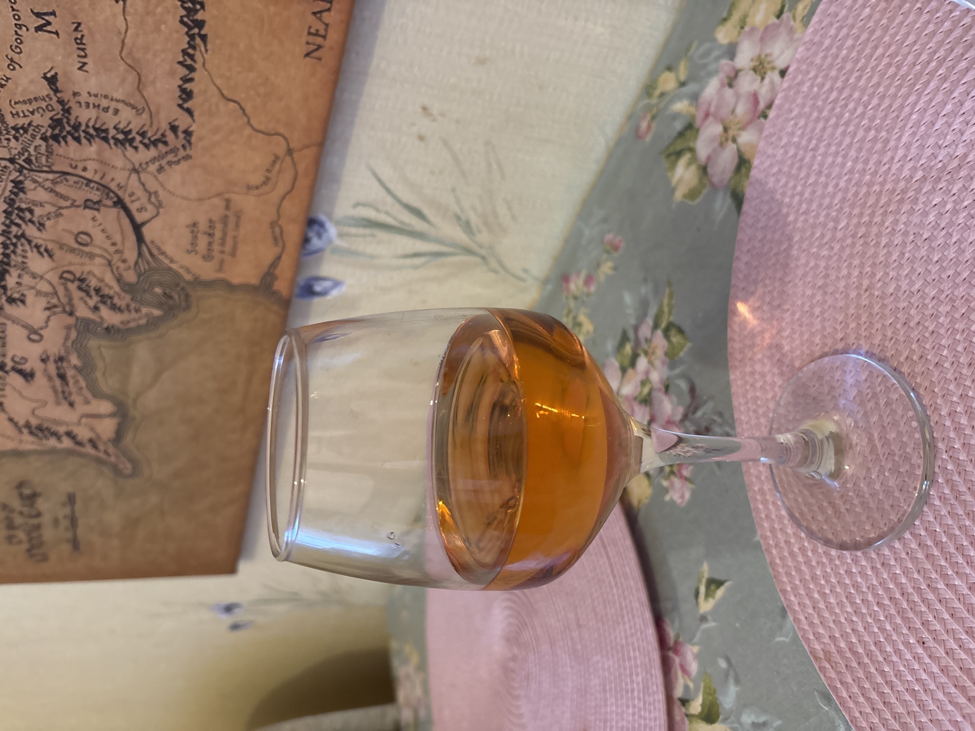 A close-up, eye-level shot focuses on a stemmed wine glass filled with amber-colored liquid, resting on a pink woven placemat. The glass is positioned to the right of the frame, its stem and base clearly visible. Behind the glass, a light blue and grey tablecloth with a floral pattern of pink and white flowers is visible. Further in the background, a map with topographical features and place names is mounted on a wall. The map appears to be a fantasy map, possibly from a fictional world. The wall behind the map has a light yellow tone with subtle floral wallpaper in shades of blue and green. No people are present in the image. The scene is indoors, likely on a table. The location is not specified, and therefore it is not possible to determine if it is in the city of Naberezhnye Chelny, Russia.